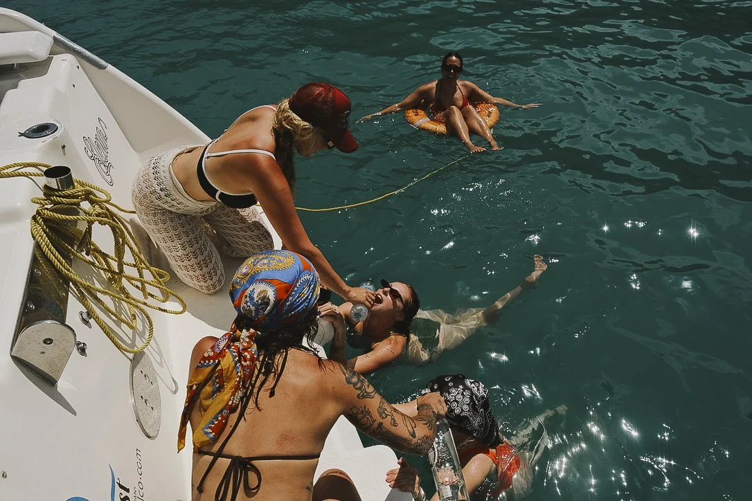 Four women on a boat assisting a woman in the water to drink water from a bottle, while another woman sits in a float in the water.