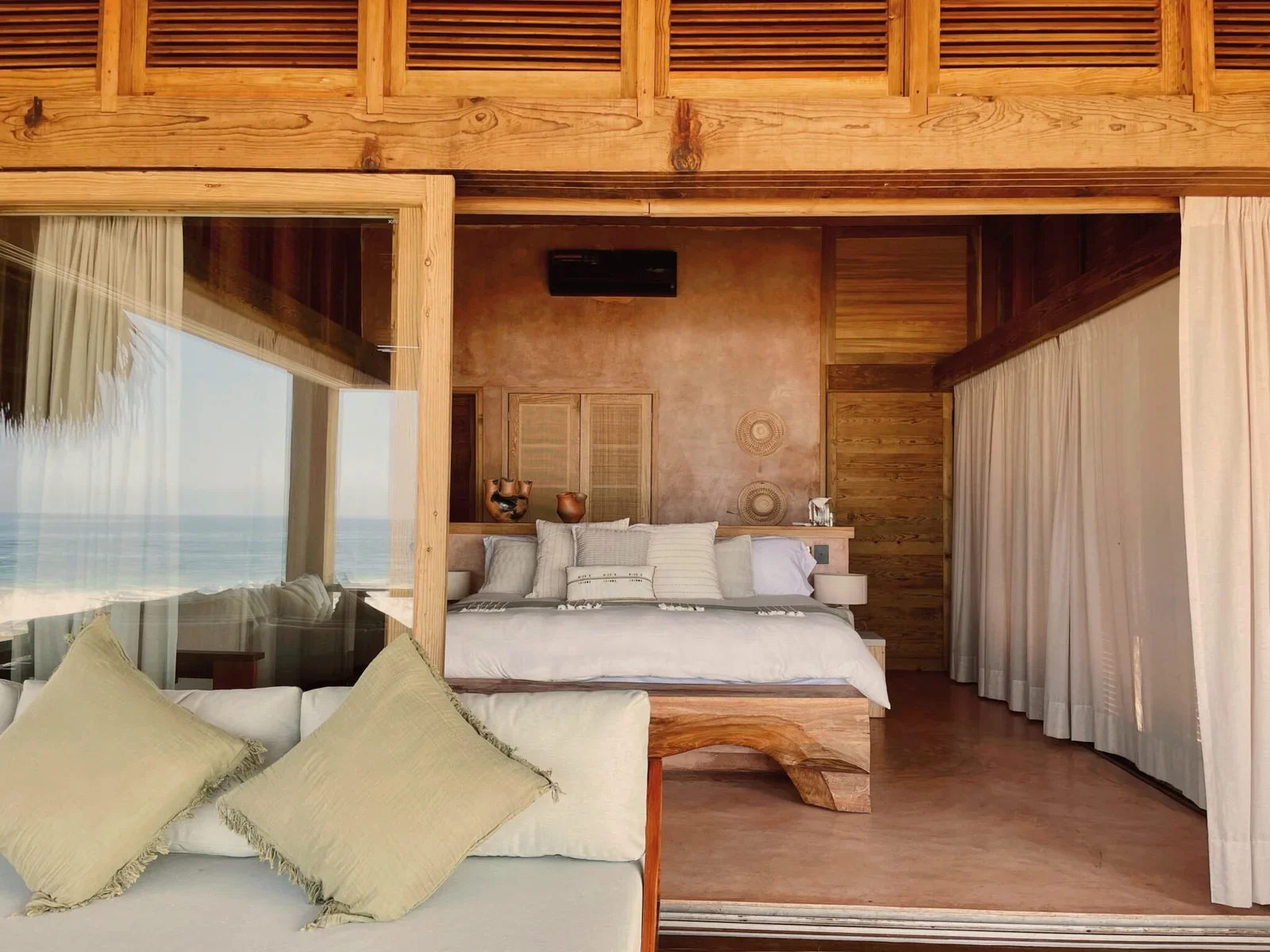 A cozy bedroom with wooden accents, a large bed with white linens, decorative pillows, and a view of the sea through a glass wall. Curtains are on the right side.