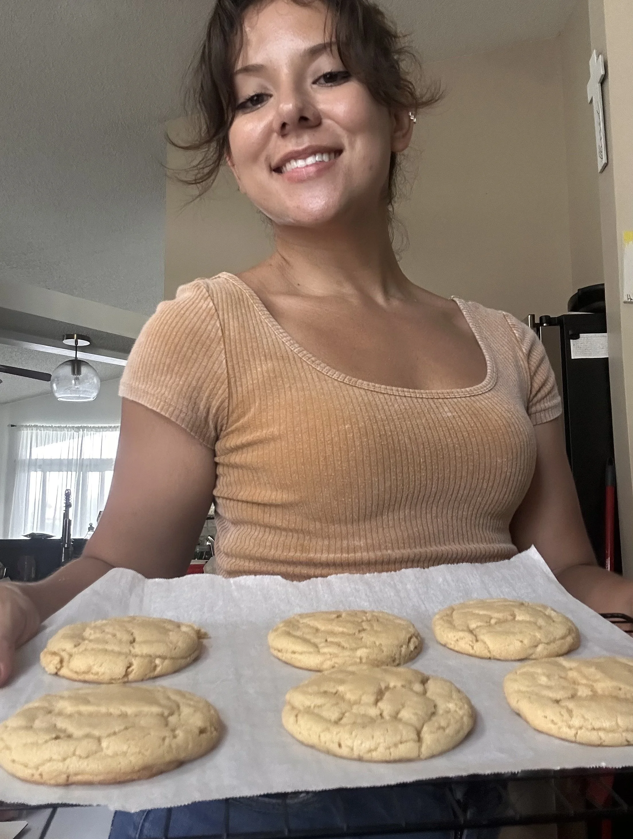 A woman with short, wavy brown hair smiles at the camera while standing behind a tray of freshly baked cookies on parchment paper. The cookies are round and golden, with a cracked surface.