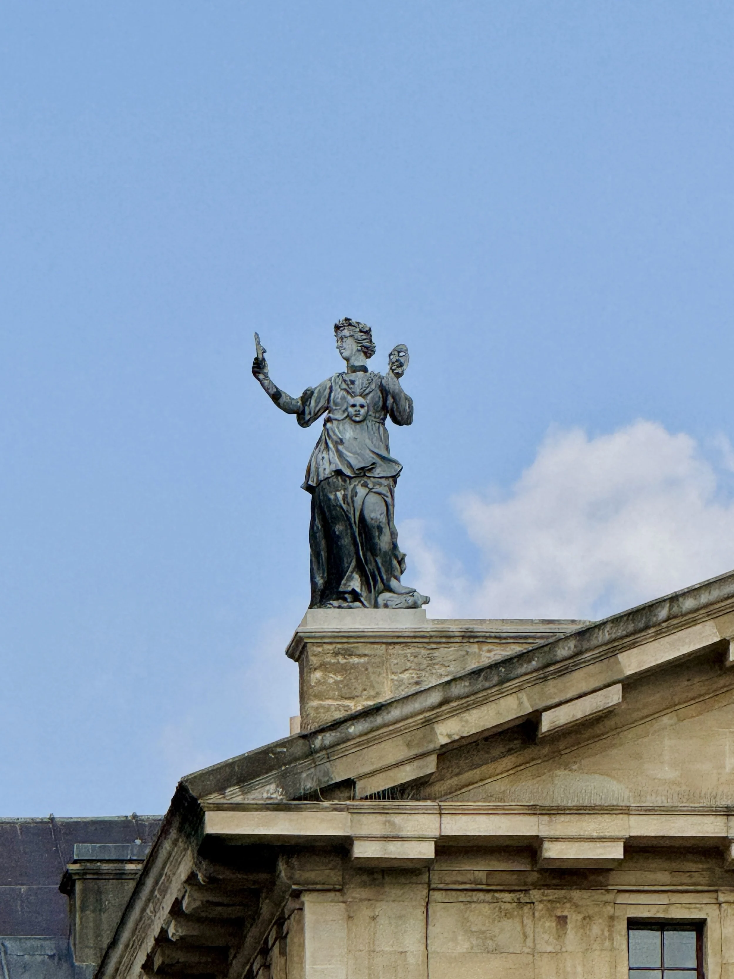 A classical stone statue of a woman holding a mask in one hand and a sword in the other, standing on the edge of a building with a blue sky in the background.