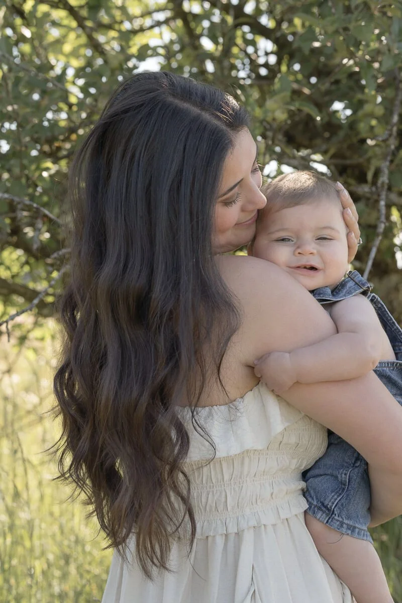 Family photographer in Olympia Washington - A woman holding a smiling young child outdoors with trees and sunlight in the background. - Angela River Photography