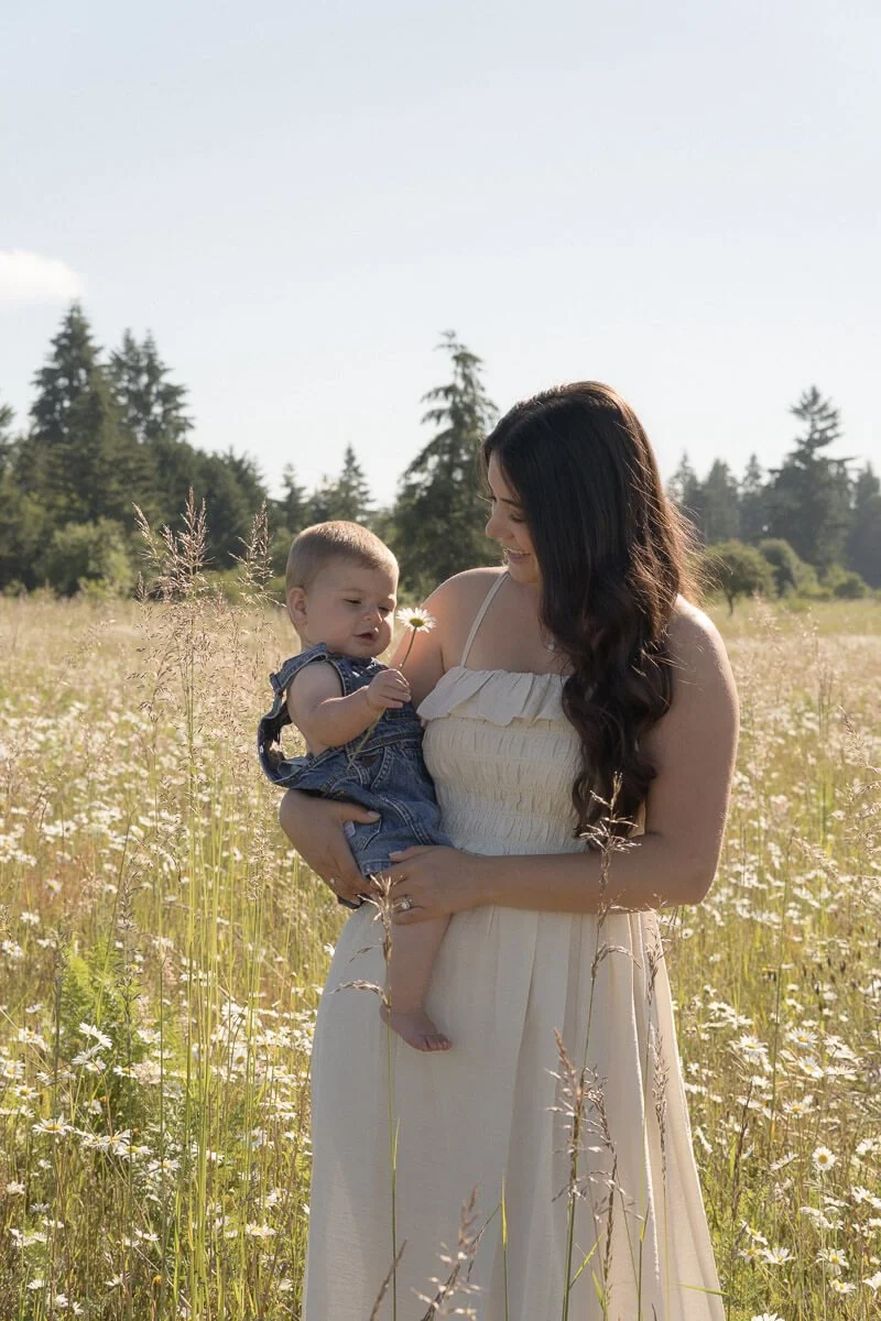 Motherhood photographer in Olympia Washington - A woman holding a baby in a field of tall grass and white flowers, with trees in the background and a clear sky overhead.- Angela Rivers Photography