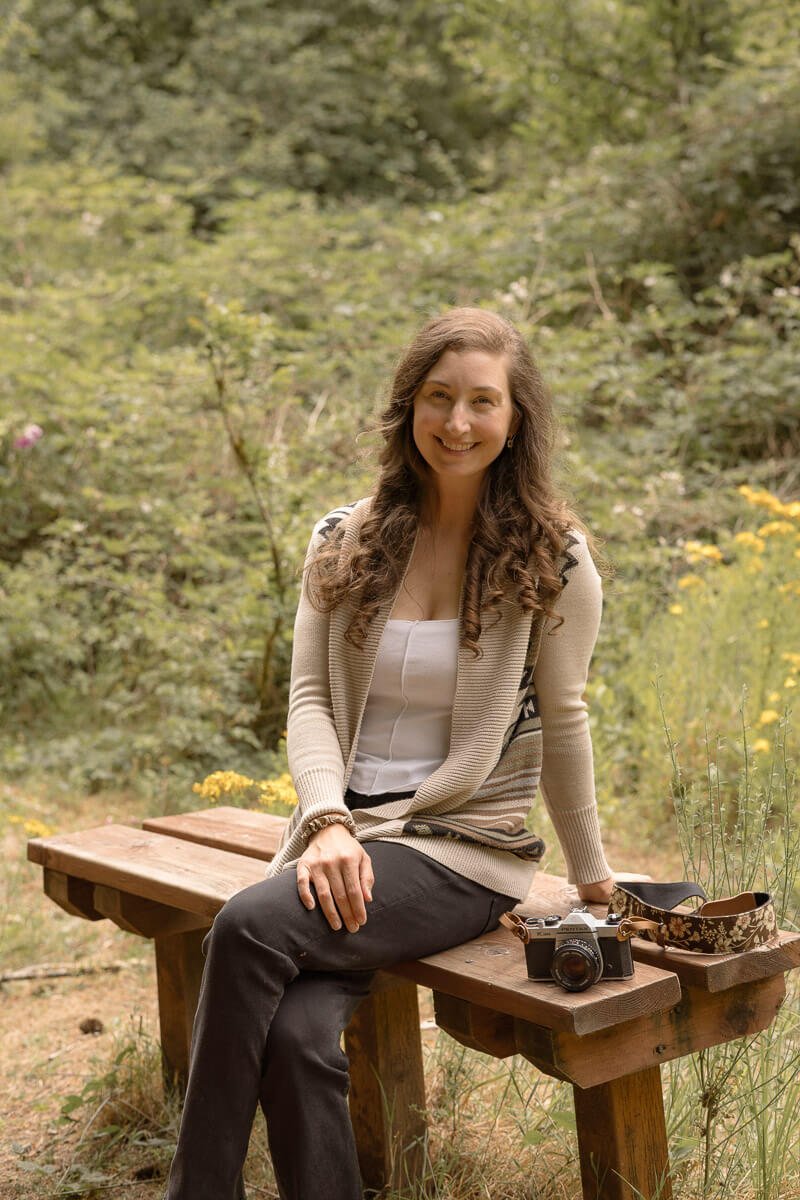 Angela Rivers — family photographer in Olympia Washington - A woman with long, curly brown hair sitting on a wooden bench in a forest, smiling at the camera. There is a camera and a camera strap on the bench next to her.