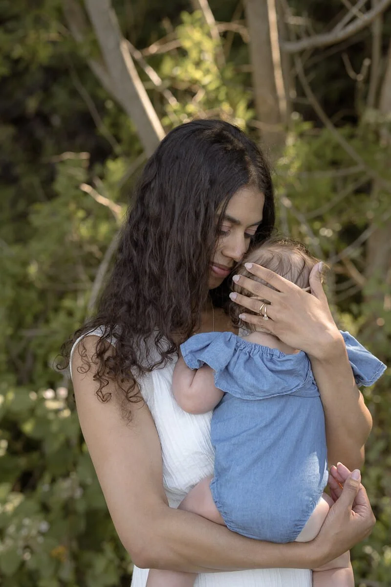 Family photographer in Olympia Washington - A woman with long, curly dark hair holding a small child close to her chest outdoors, surrounded by trees and greenery. - Angela Rivers Photography