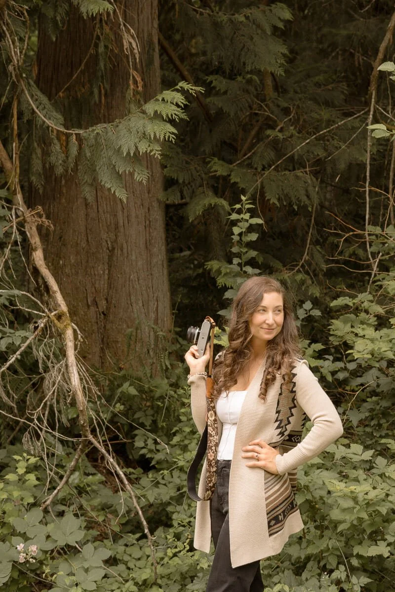 Angela Rivers — family photographer in Olympia Washington - A woman standing in a lush forest holding a camera in her right hand, wearing a beige and patterned cardigan, with long wavy hair and a slight smile.