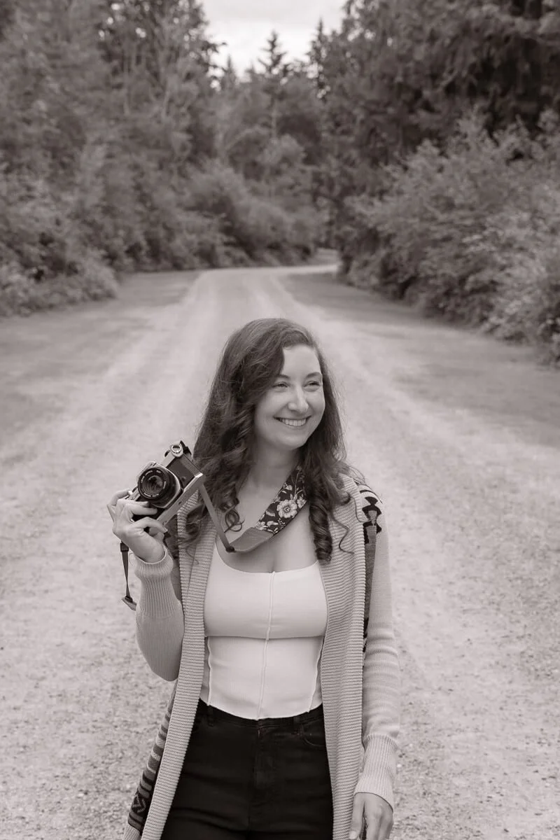 Angela Rivers — family photographer in Olympia Washington - A woman smiling and holding a camera, standing on a dirt road surrounded by trees.