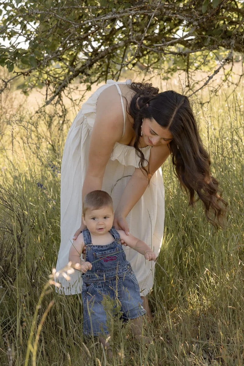 Documentary family photographer in Olympia Washington - A woman in a white dress crouches down to help a young child who is standing in tall grass in a park or meadow, with trees in the background. - Angela Rivers Photography