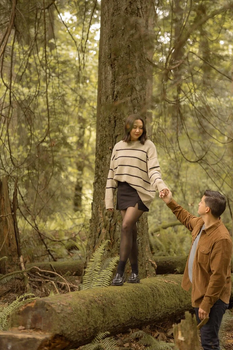 Engagement photographer in Olympia Washington - A woman in a beige and black striped sweater and black skirt standing on a fallen tree in a forest, holding hands with a man in a brown jacket and black pants. - Angela Rivers Photography