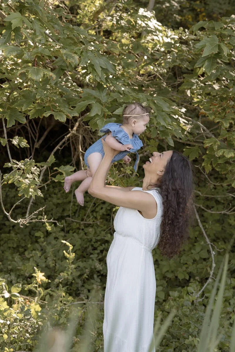 Mom and baby photographer in Olympia WA - A woman with long dark hair lifting a smiling baby girl in a blue dress in a lush green outdoor setting. - Angela Rivers Photography