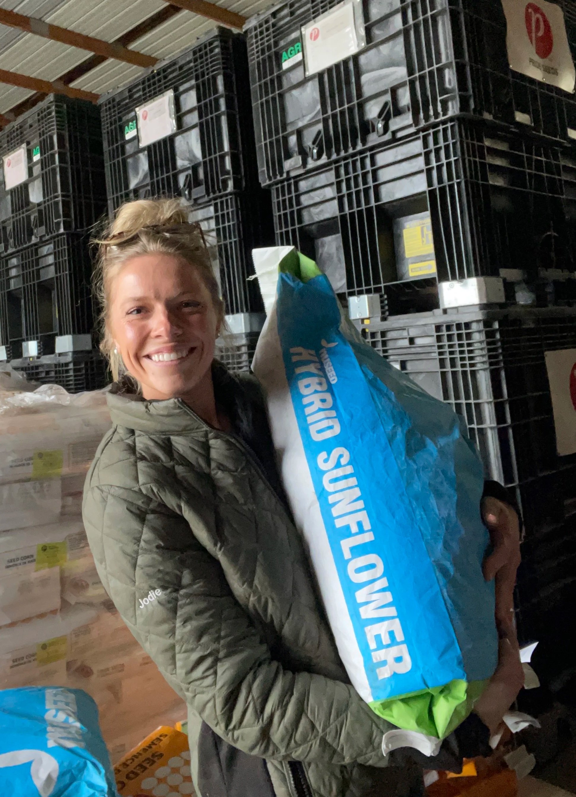 A woman with blonde hair smiling, wearing a green jacket, holding a large blue and white bag of Sunflower Seeds. She is standing in front of stacked crates and boxes in a warehouse or storage area.
