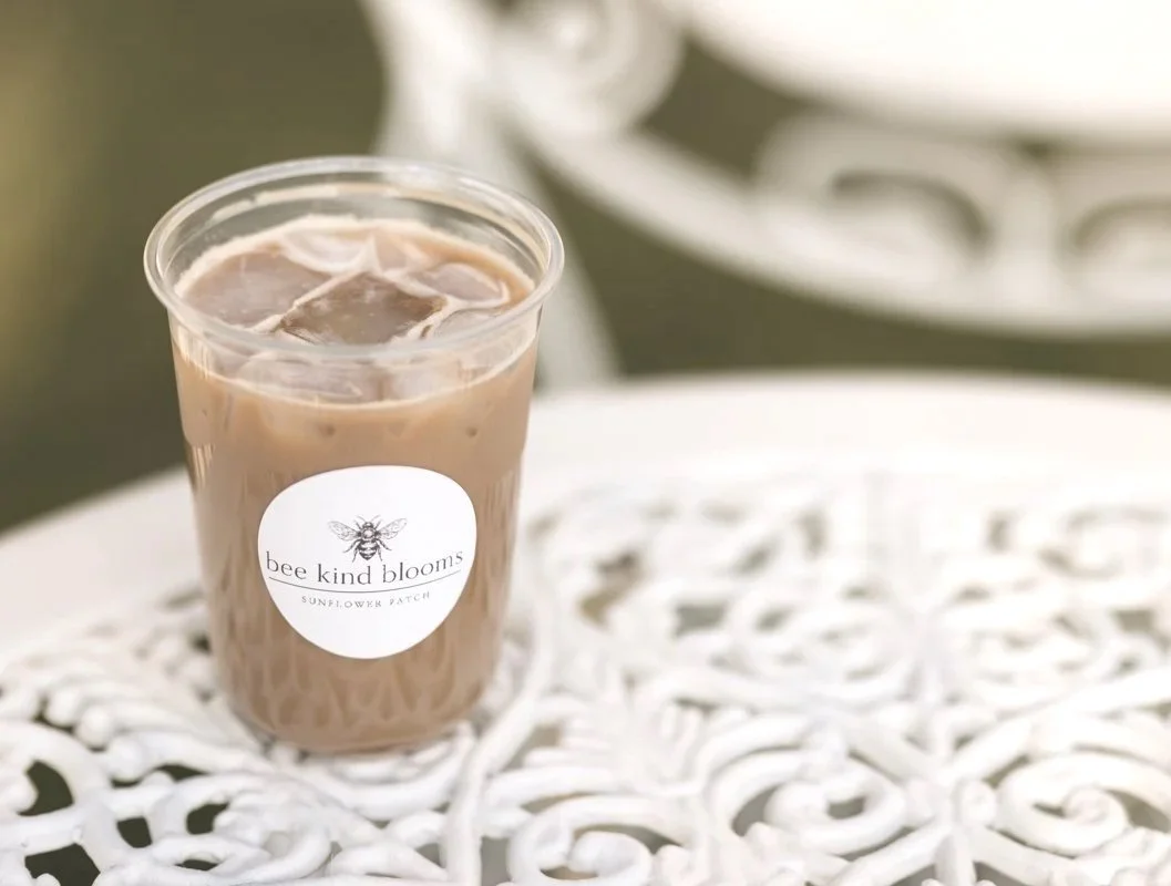 A plastic cup filled with iced coffee on a white ornate metal table, with a sticker reading "bee kind blooms" and a bee logo.