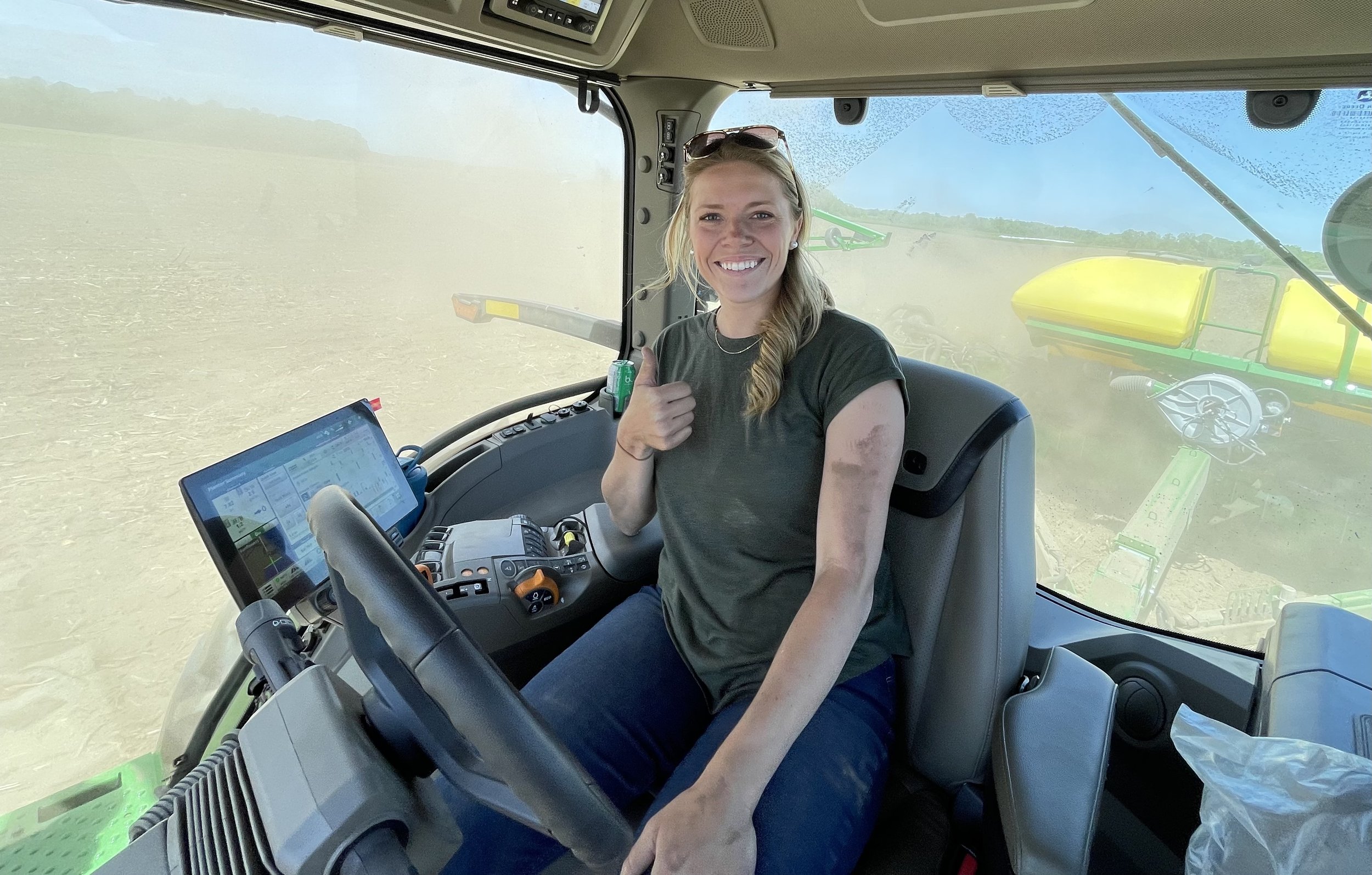 Smiling woman wearing sunglasses inside the cabin of a tractor, sitting at the controls, with a large landscape of field and blue sky visible through the window behind her.