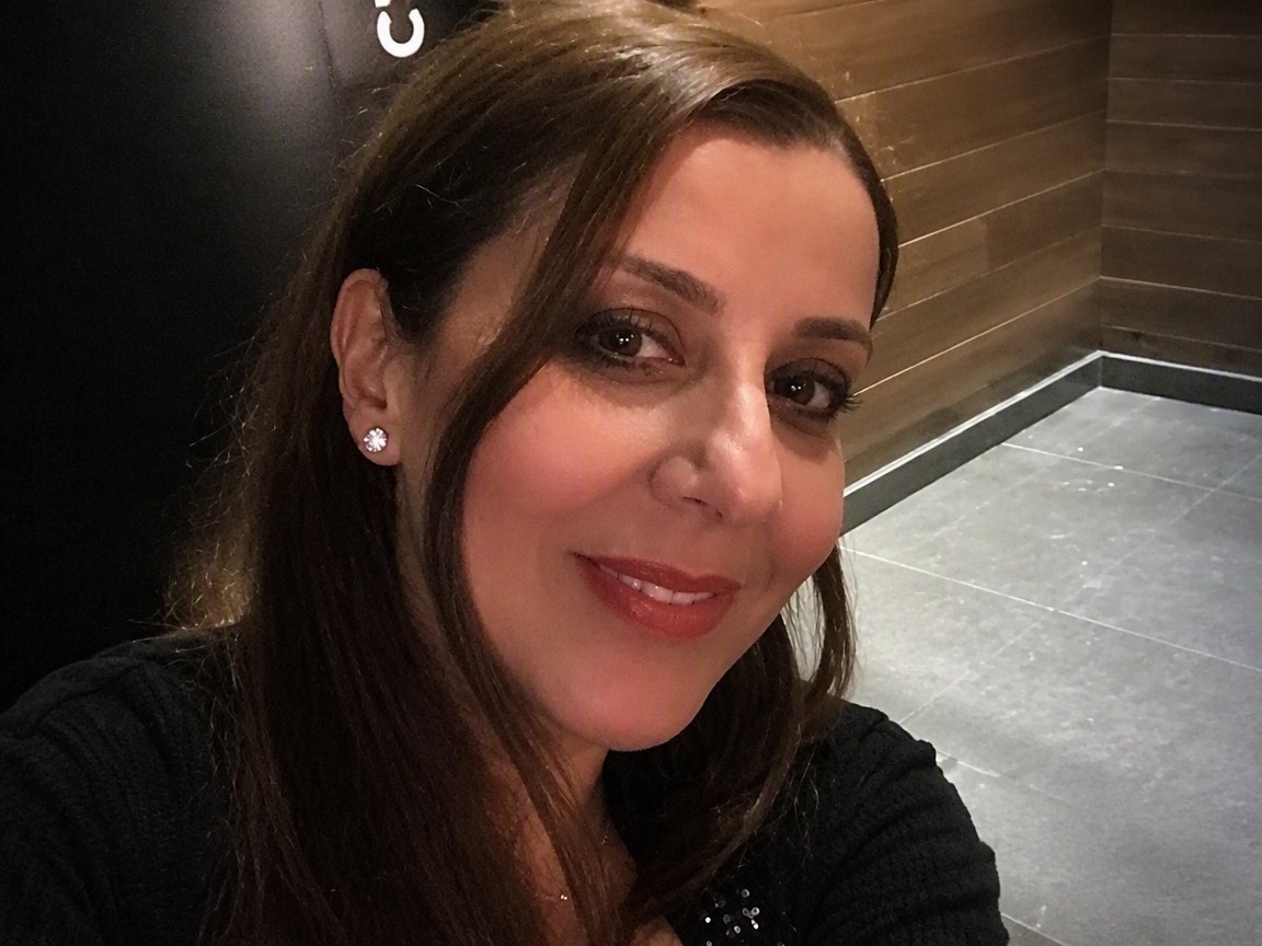 Une femme souriante avec des boucles d'oreilles en forme de fleur, portant un maquillage léger, dans un intérieur moderne avec un mur en bois et un sol en carrelage gris.