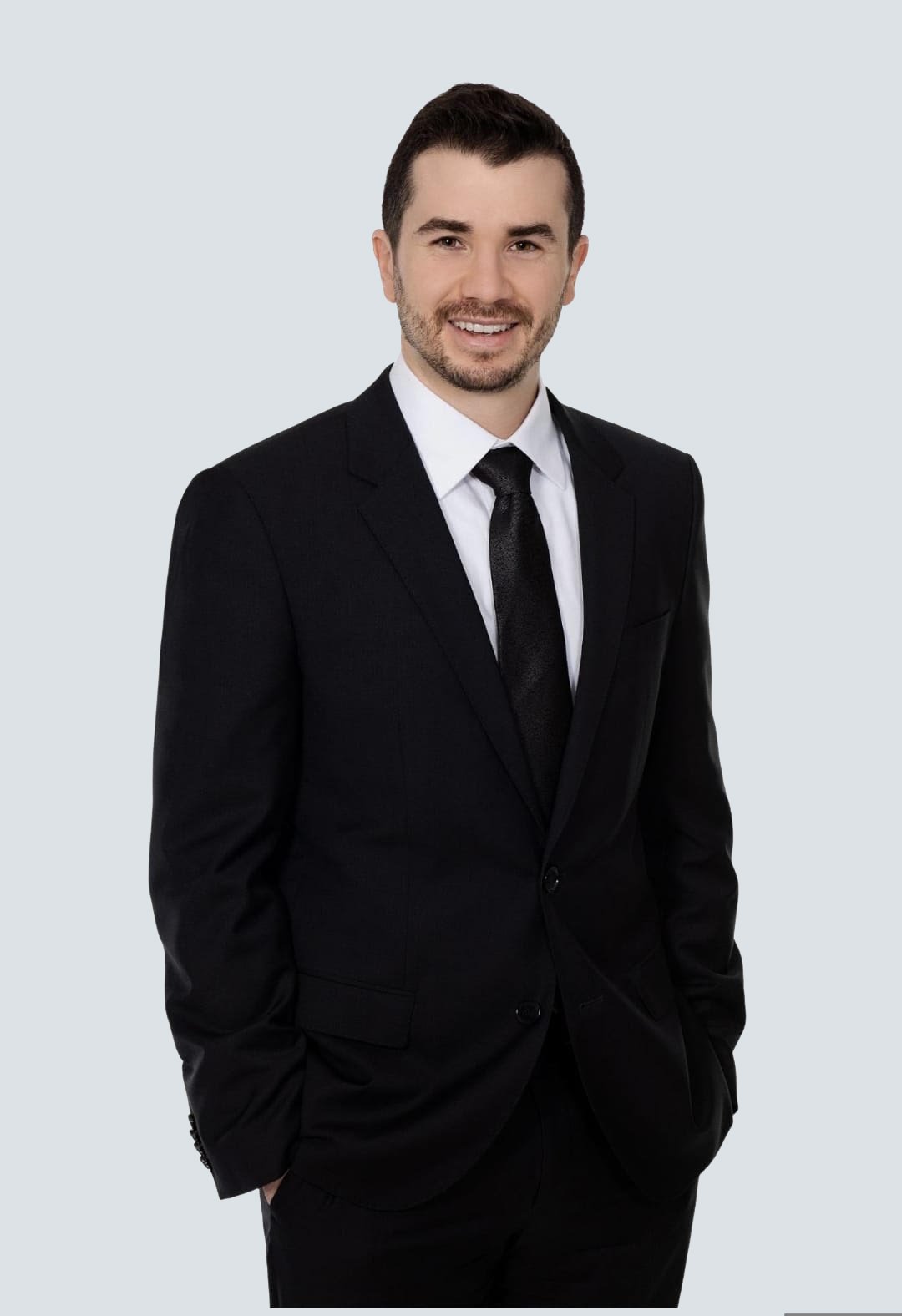 Homme d'affaires en costume noir avec une cravate noire, souriant, posant contre un fond gris clair.