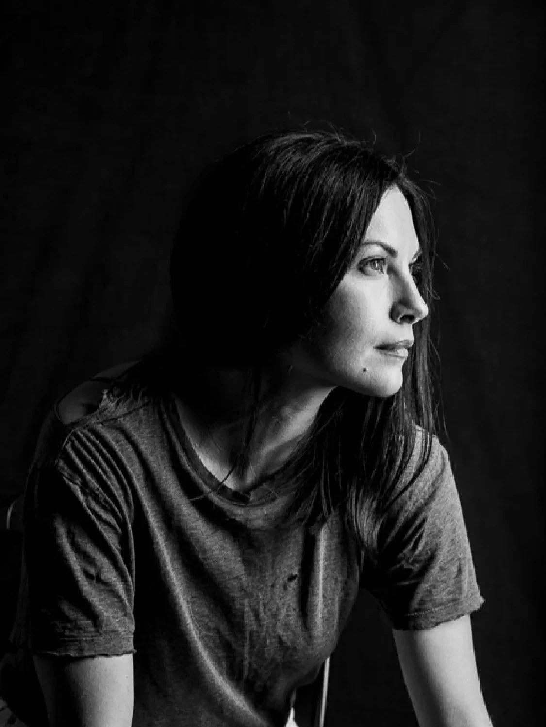 A black and white portrait of a woman with shoulder-length dark hair, looking to the right with a thoughtful expression, wearing a casual gray t-shirt.
