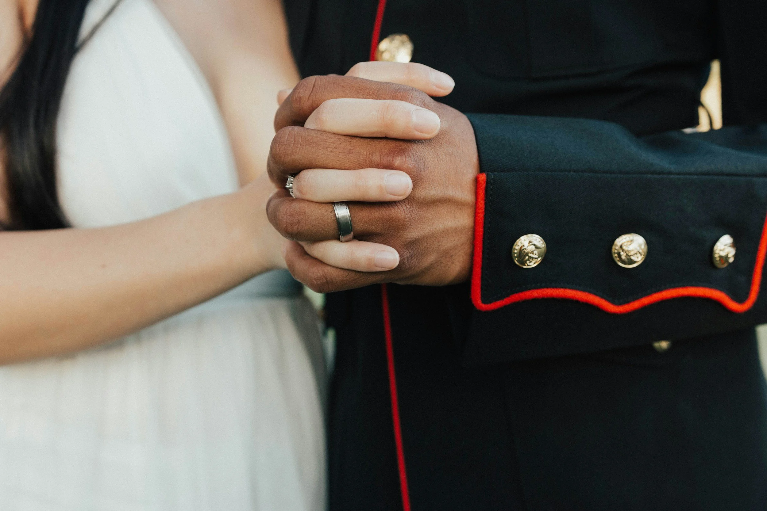 Close-up of two people clasping hands, one in a wedding ring, with one person wearing a military uniform with gold buttons and red piping.