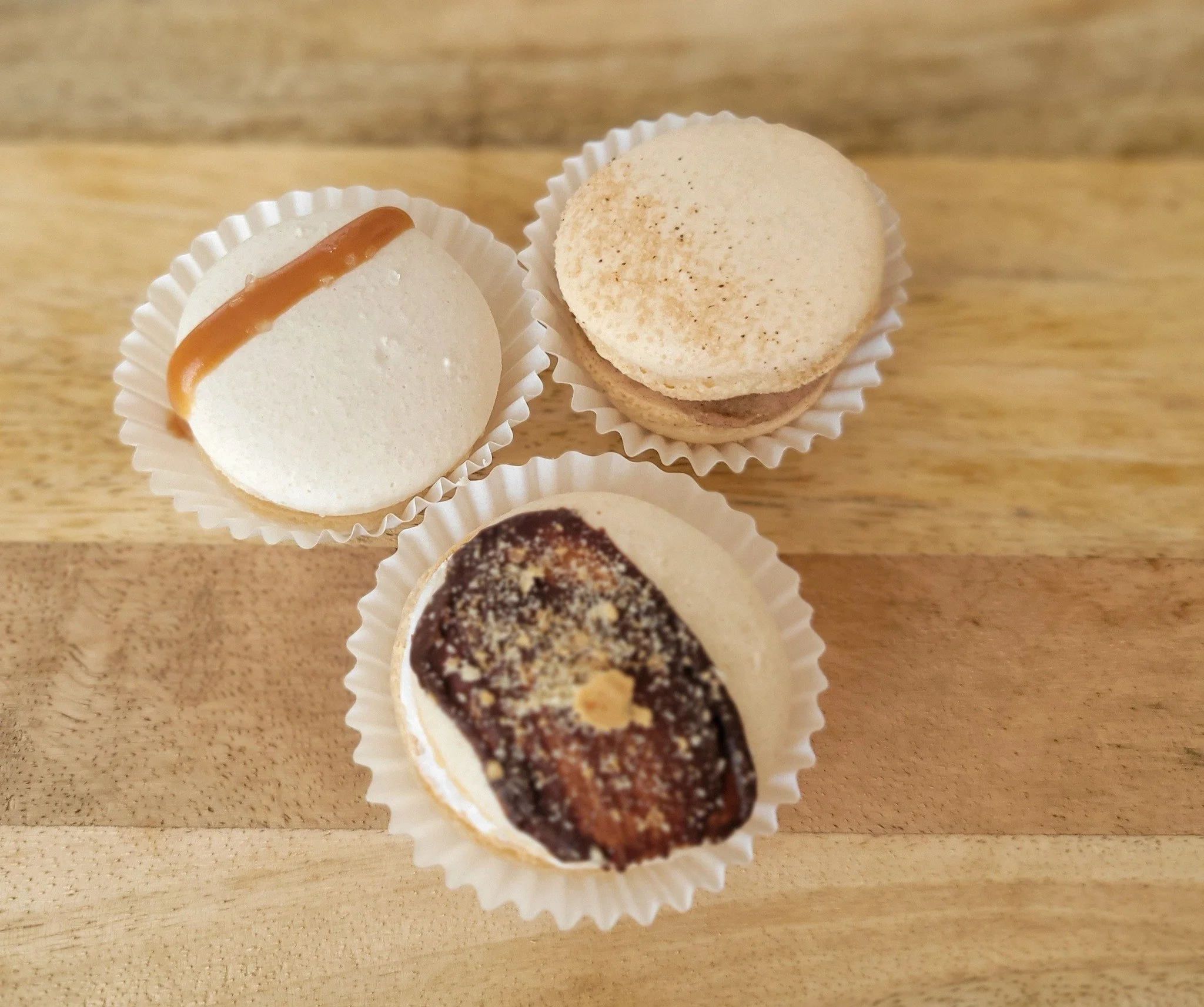 Close-up of three small round confections in white paper cups on a wooden surface, featuring creamy white, light brown, and dark chocolate with chopped nuts toppings.