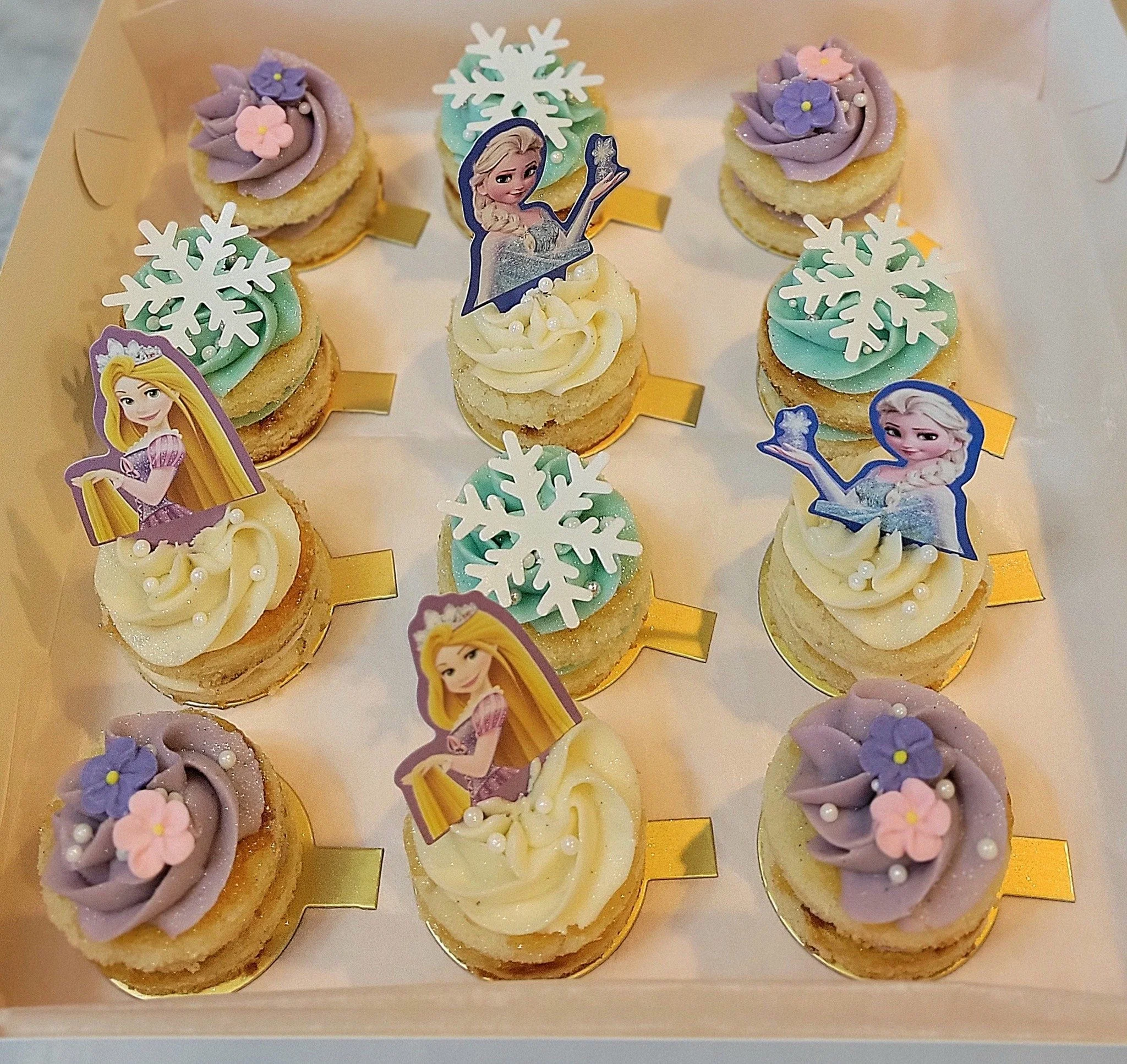 A box of cupcakes decorated with princess and snowflake themed toppers, with purple and pink frosting, pearls, and edible flowers.