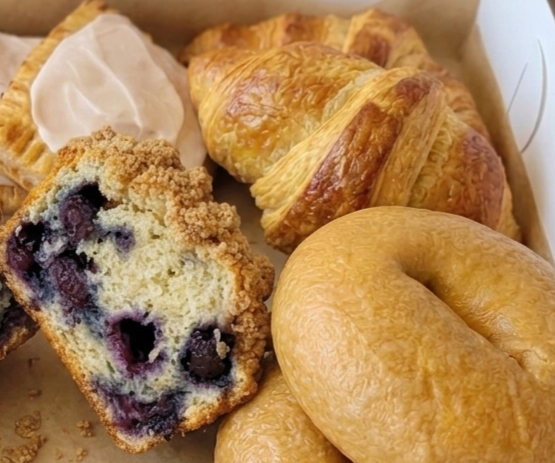 Assorted baked goods including a blueberry muffin, a croissant, a bagel, and a muffin with icing, in a bakery box.