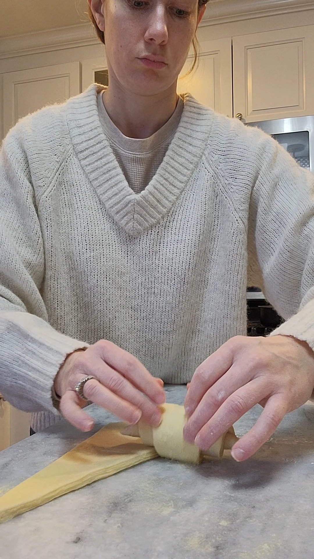 A person wearing a cream-colored knitted sweater is rolling out dough on a marble countertop in a kitchen.