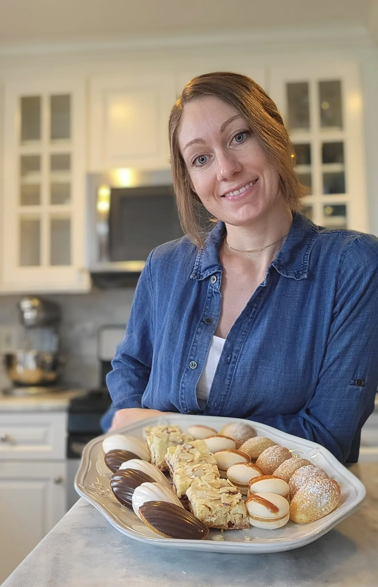 A woman with short brown hair and a denim shirt standing behind a large platter of assorted cookies and desserts in a kitchen.