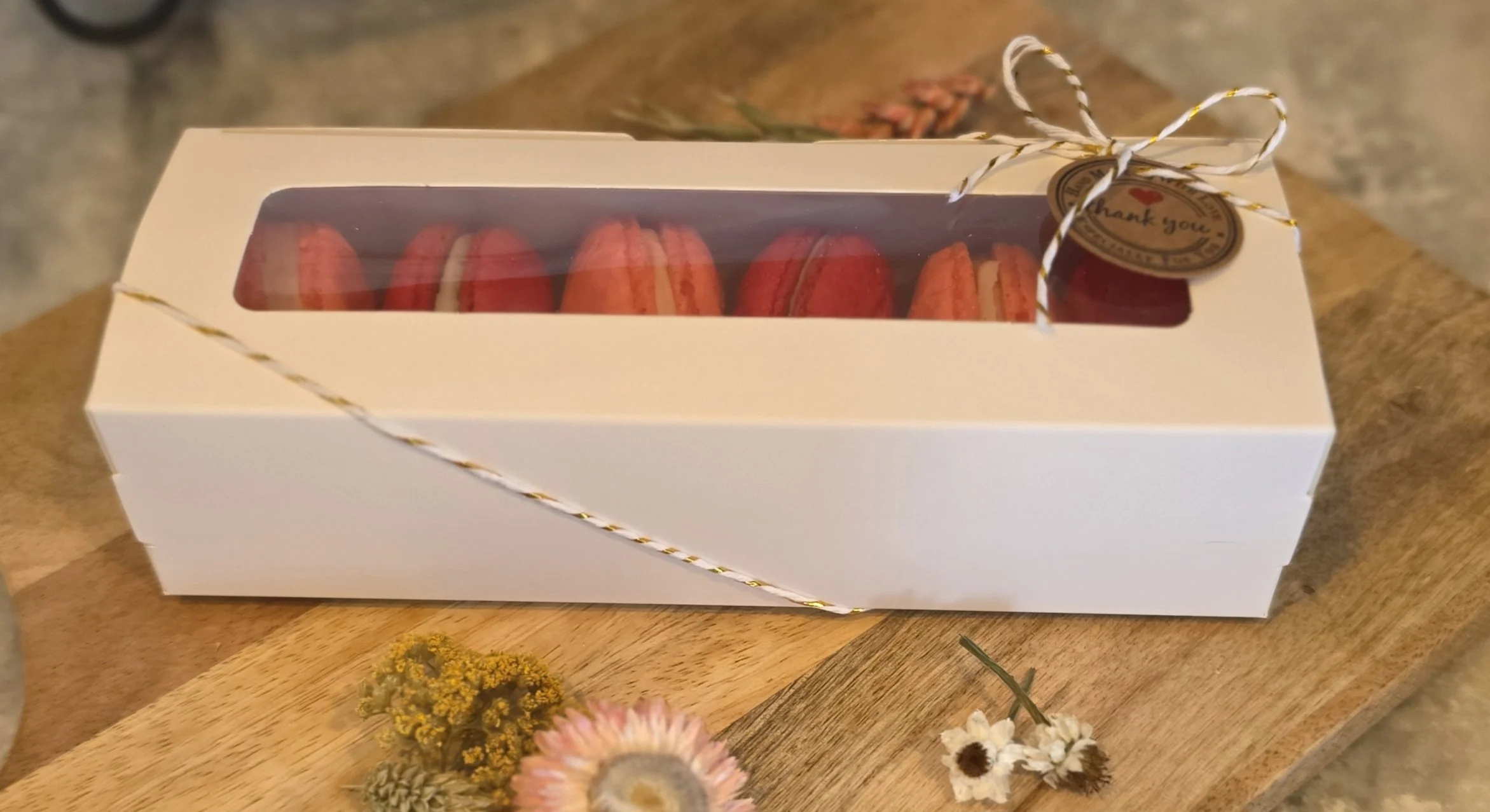 A white gift box with a clear top revealing red and white cookies inside, decorated with gold and white twine and a 'thank you' sticker, placed on a wooden surface surrounded by small colorful flowers.