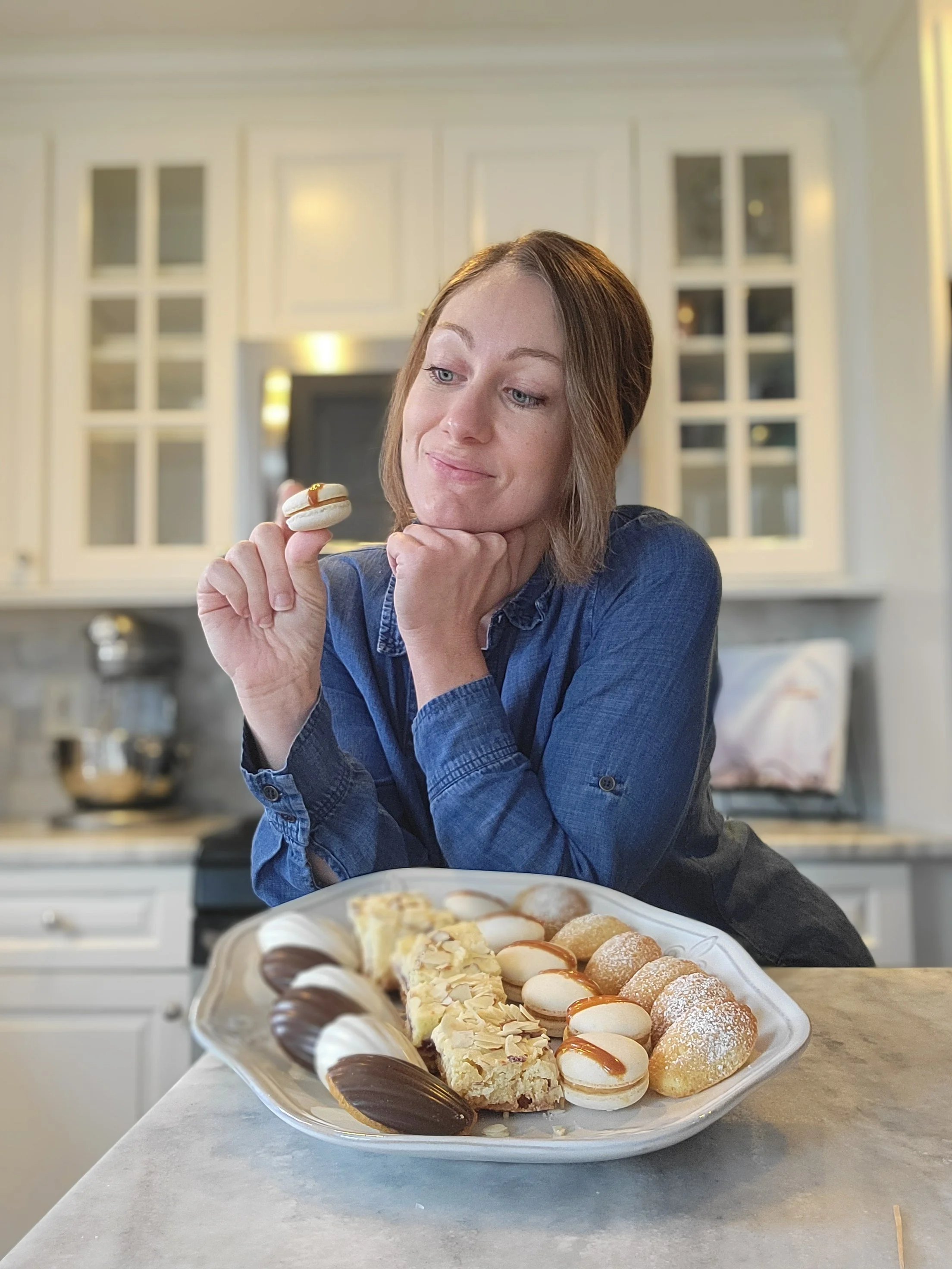 A woman is sitting at a kitchen counter and holding a macaron in her right hand. She is looking at the cookie with a slight smile, resting her chin on her left hand. The counter holds a tray of assorted cookies and desserts.