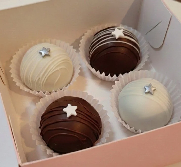 Four decorated chocolates in a pink box, with two white and two dark chocolate pieces, each topped with a small silver or white star decoration.