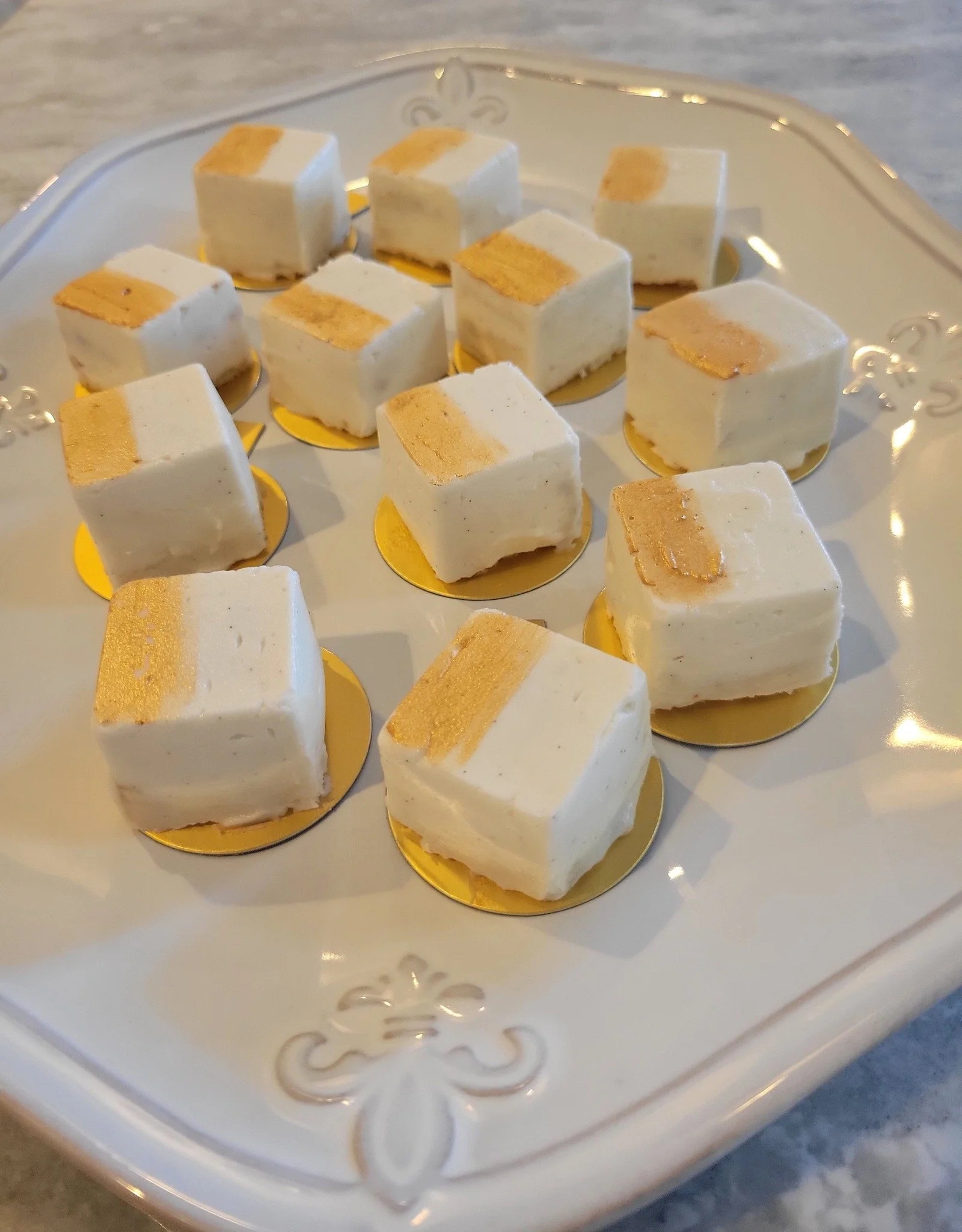 A white decorative tray with gold accents holds small square cakes with white frosting and a gold stripe on top.