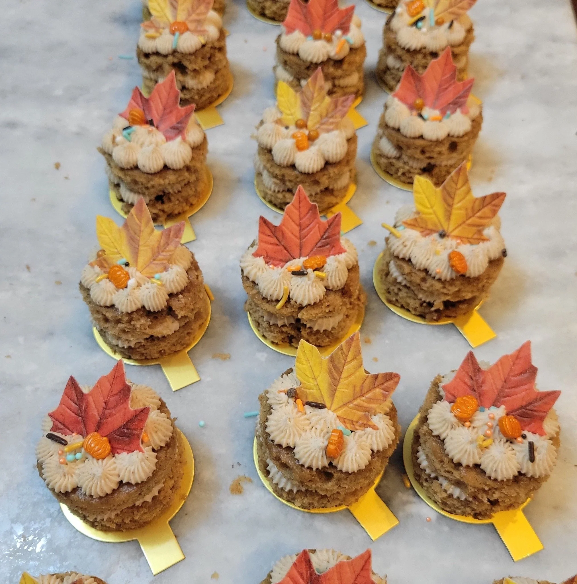 A display of mini layers decorated with piped icing, orange sprinkles, and fall leaves, arranged on small yellow cardboard bases on a marble surface.