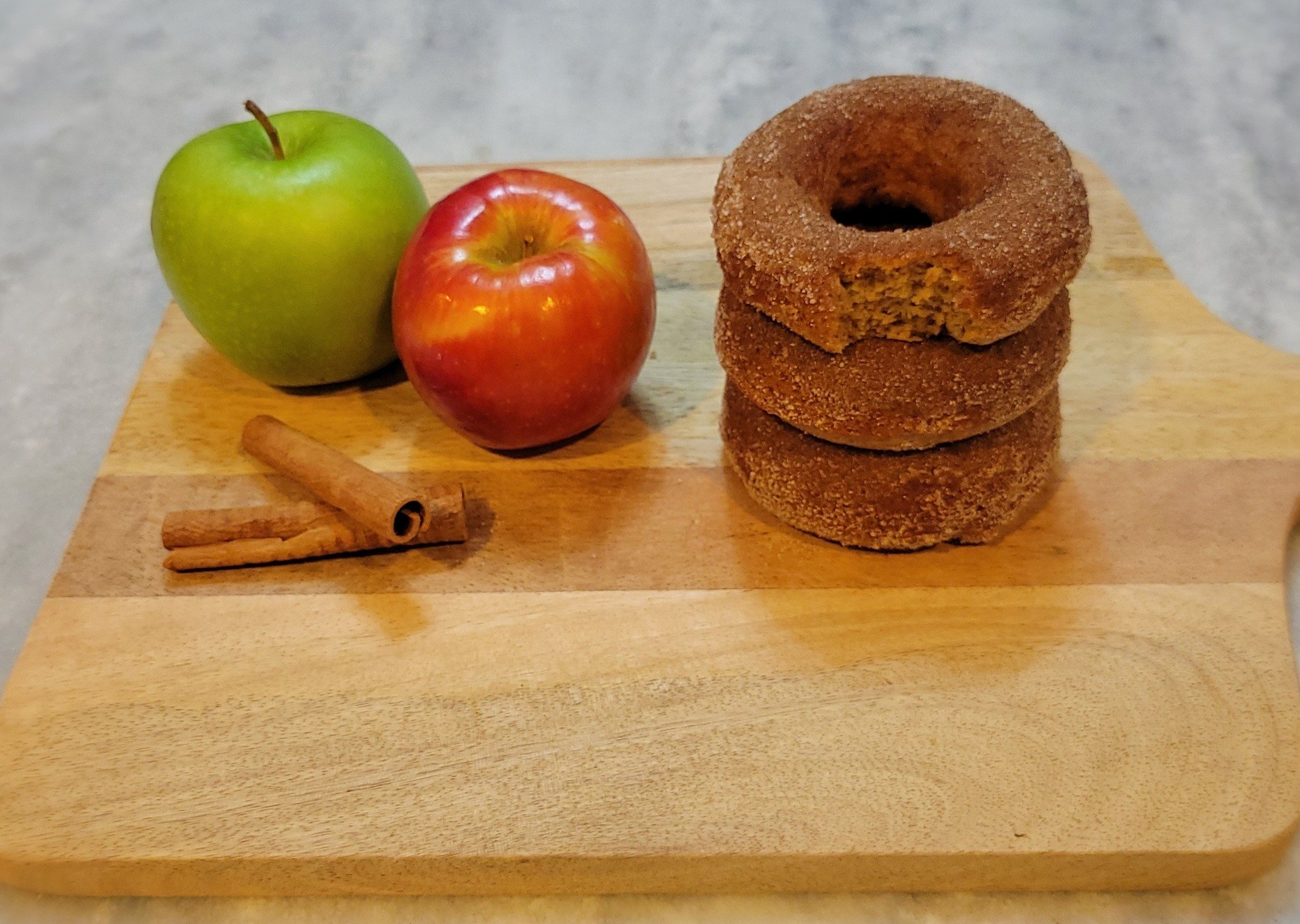 Two apples, one green and one red, along with two cinnamon sticks, placed on a wooden surface with three stacked cinnamon donuts on the right.