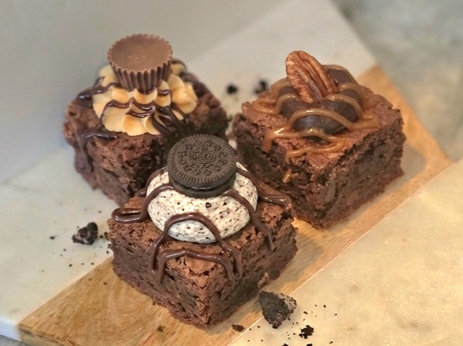 Three chocolate brownies topped with various toppings, placed on a wooden board. The first brownie has a Reese's peanut butter cup, and chocolate drizzle. The second has pecan half, and chocolate drizzle. The third has an Oreo cookie, and chocolate d