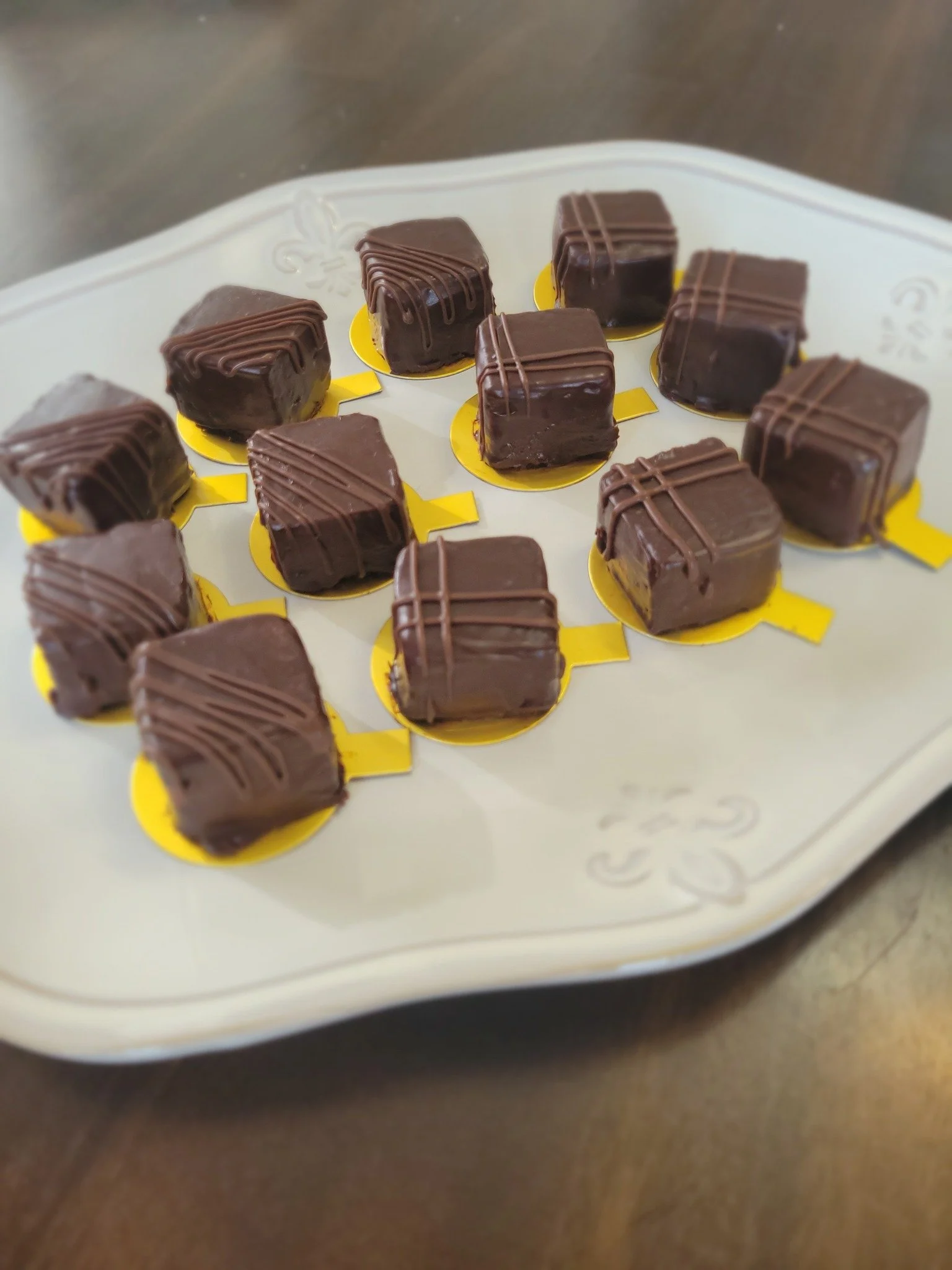 Close-up of small square-shaped chocolates with a glossy chocolate coating and decorative drizzle, arranged on a white tray with a gold border.