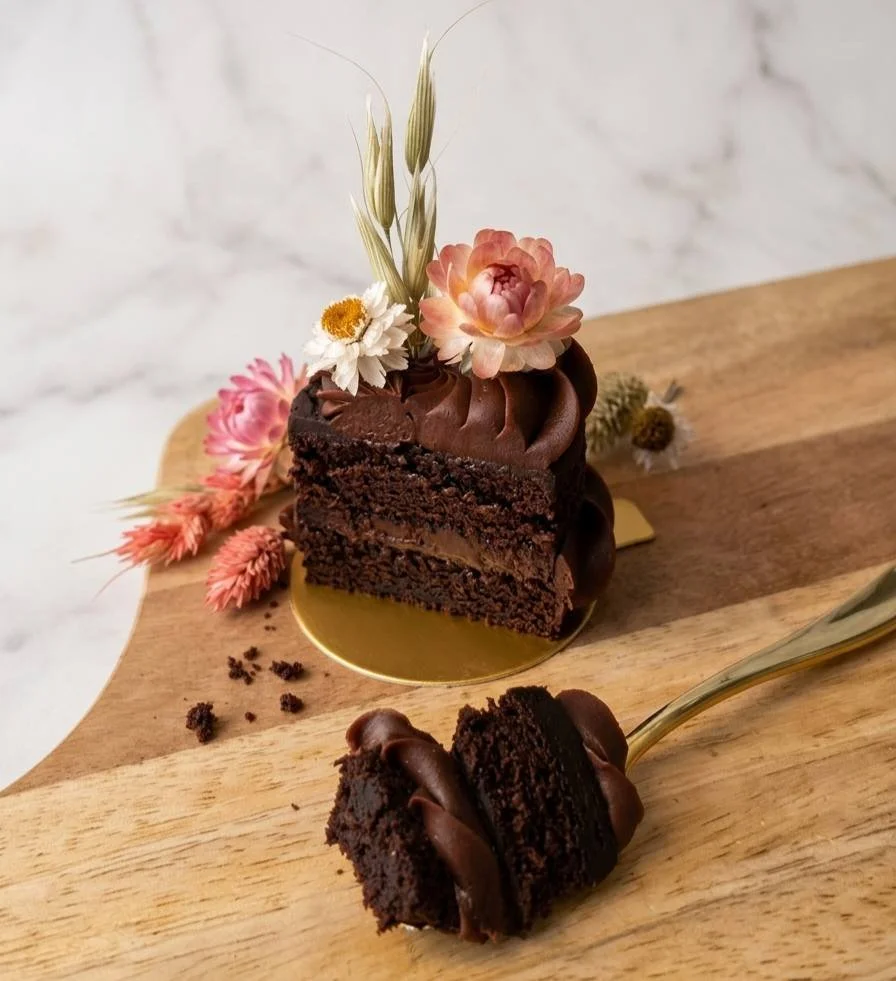 A slice of chocolate cake with frosting topped with pink, white, and orange flowers, placed on a gold cake board on a wooden board, with a fork holding a piece of cake.