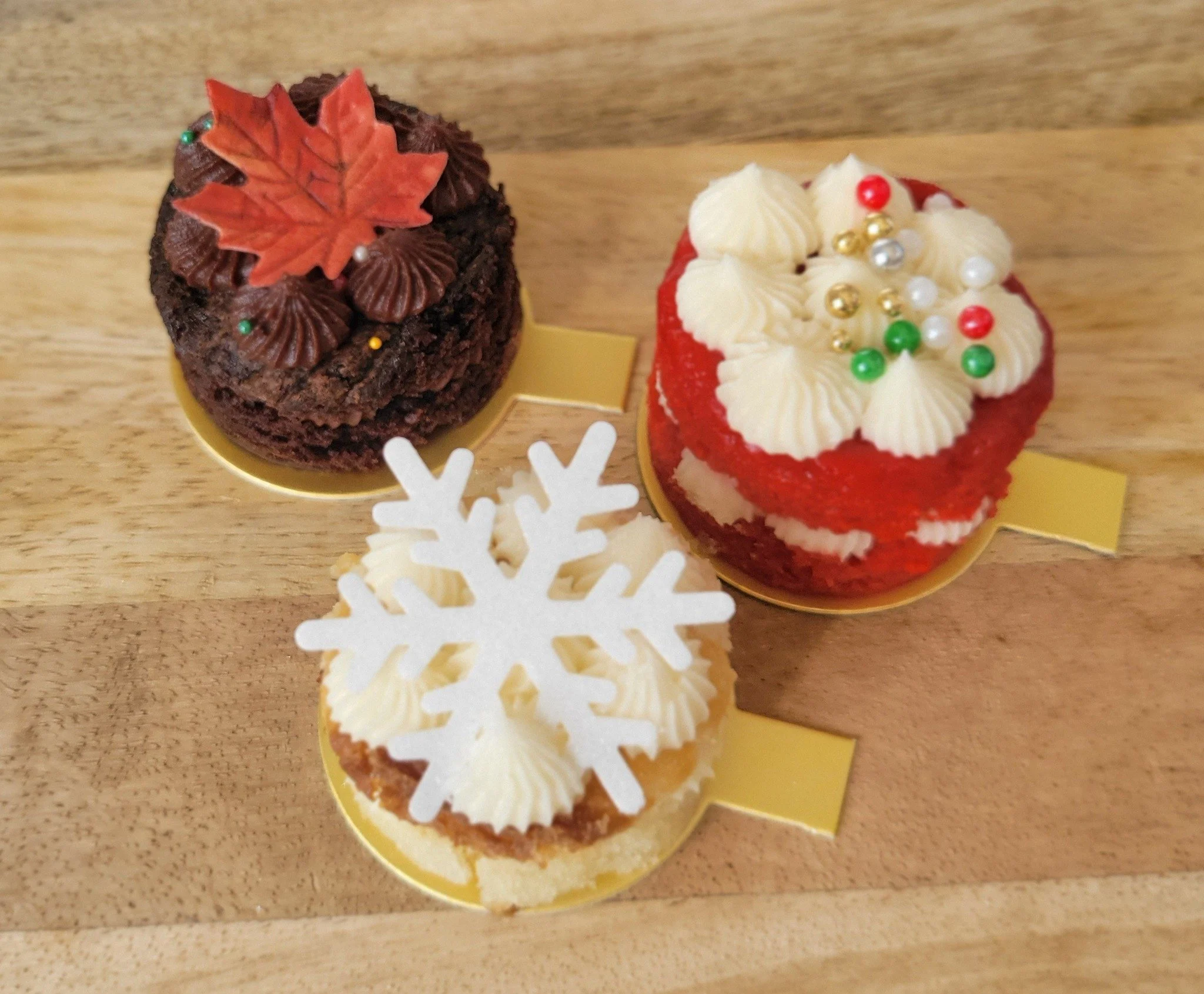 Chocolate cupcake decorated with a red maple leaf, small colored sprinkles, and green sugar beads; vanilla cupcake decorated with white frosting, small colored sprinkles, and a red candy on top, both placed on small gold bases on a wooden surface wit
