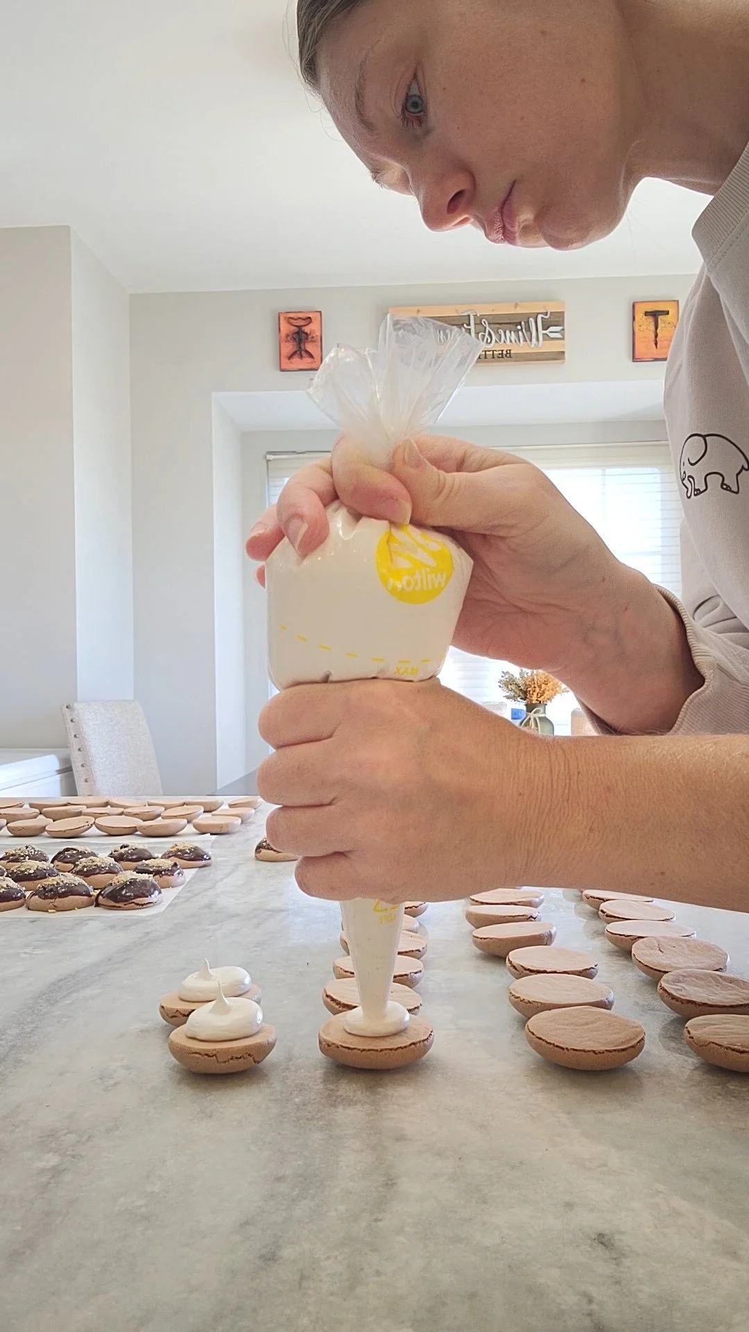 A woman decorating cookies with white icing in a kitchen with a table filled with cookies and a window in the background.