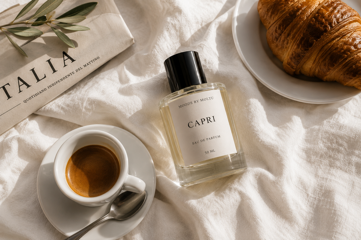 A cup of espresso on a saucer with a teaspoon, a bottle of 'Capri' perfume, a croissant on a white plate, and a newspaper with a branch of green leaves on a cream-colored fabric surface.