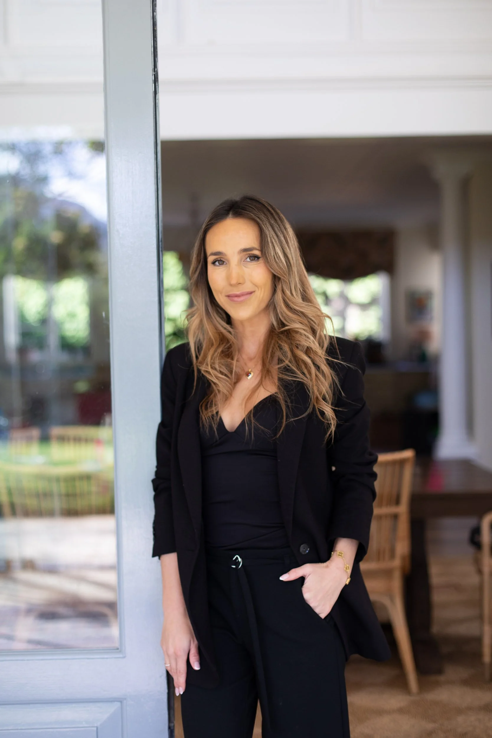 A woman with long, wavy brown hair wearing a black blazer and black pants, standing inside near a glass door with a blurred outdoor background.