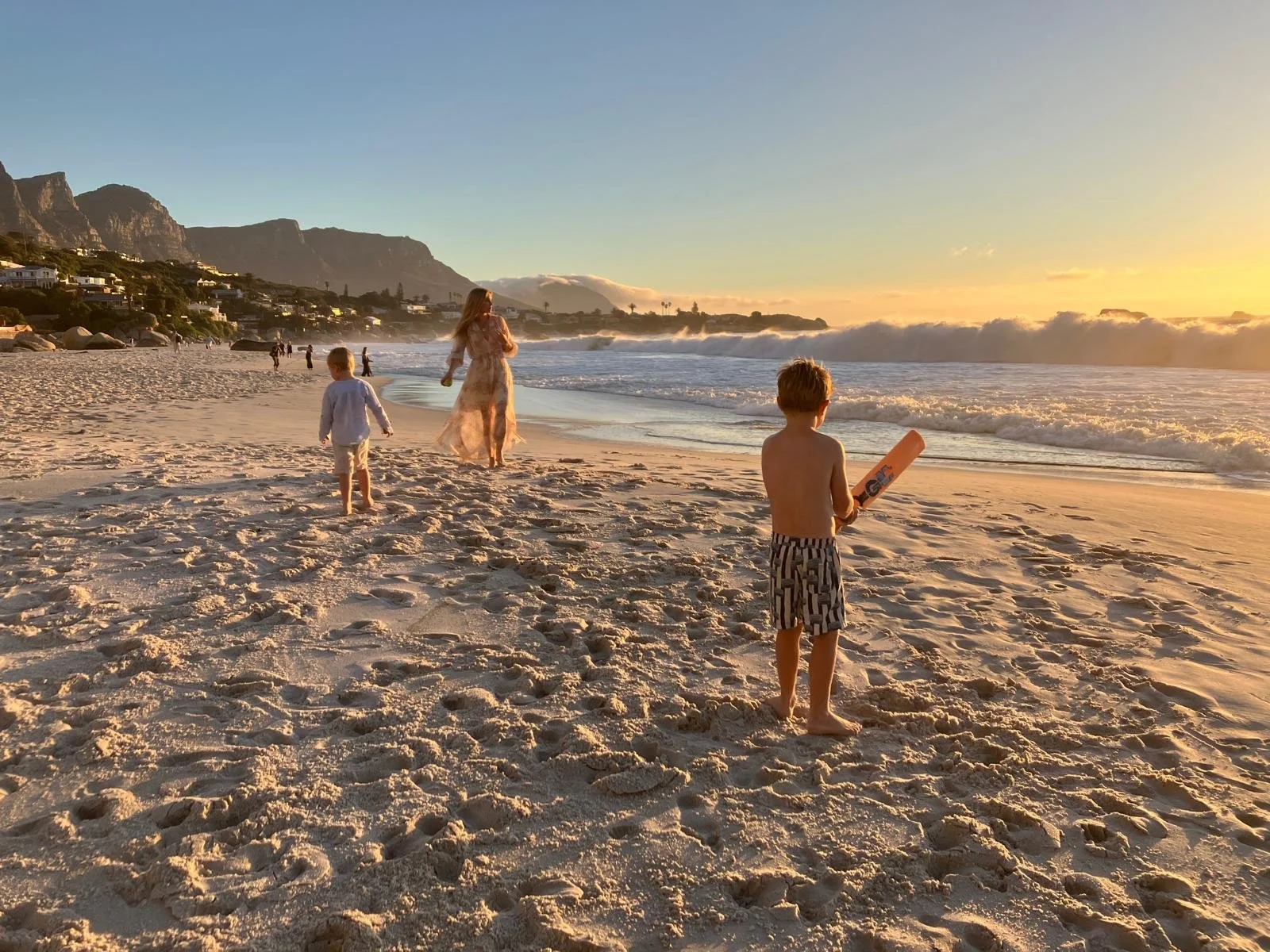 Children and a woman walking on the sandy beach at sunset with waves crashing and mountains in the background.