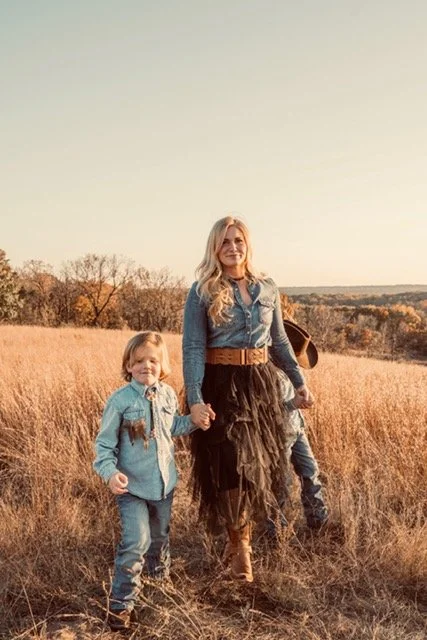 A woman and two children walking through a grassy field at sunset, holding hands, with trees and rolling hills in the background.