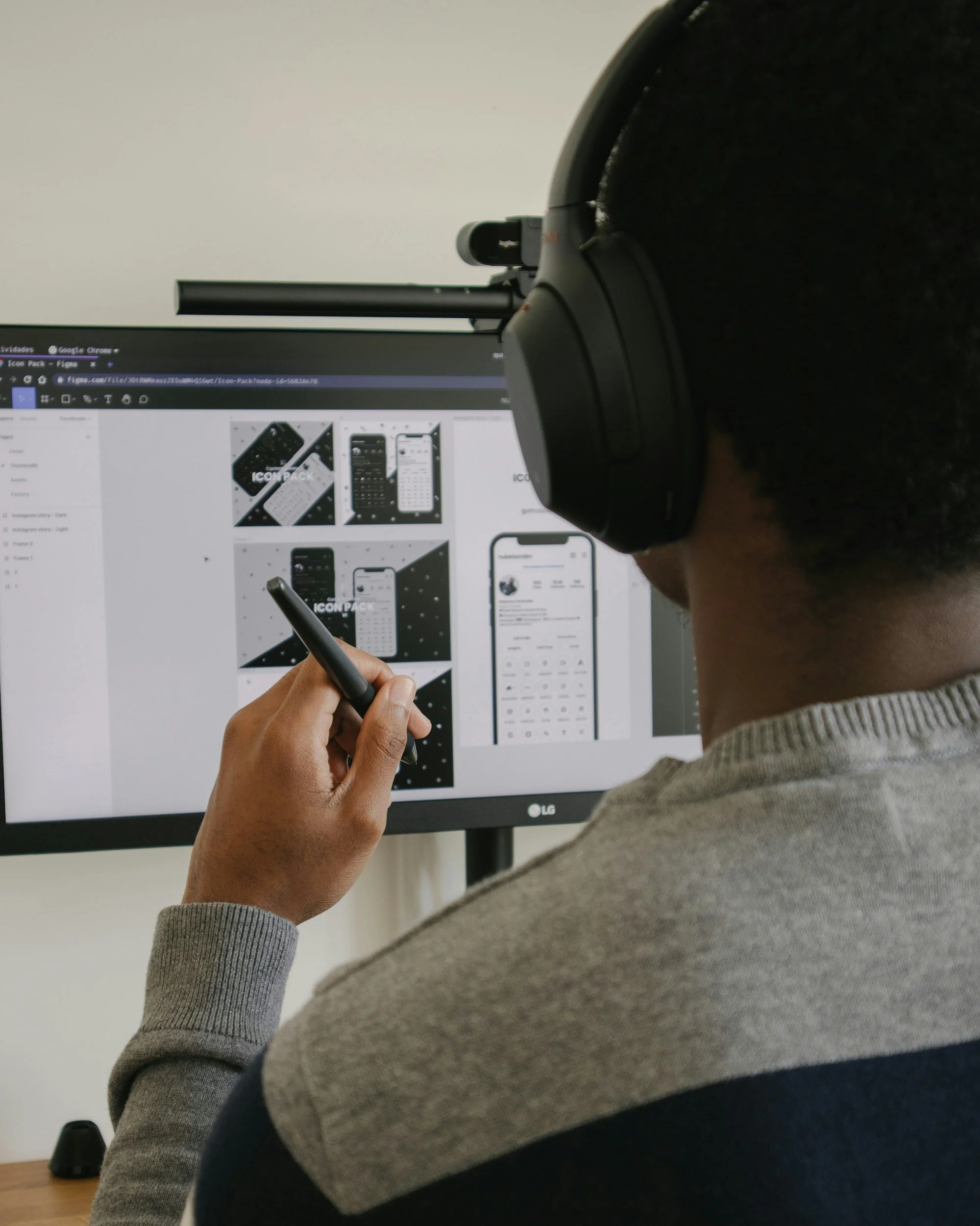 Person with dark hair wearing large over-ear headphones, sitting in front of a computer monitor displaying design concepts for a mobile app, holding a stylus pen, in a well-lit room.