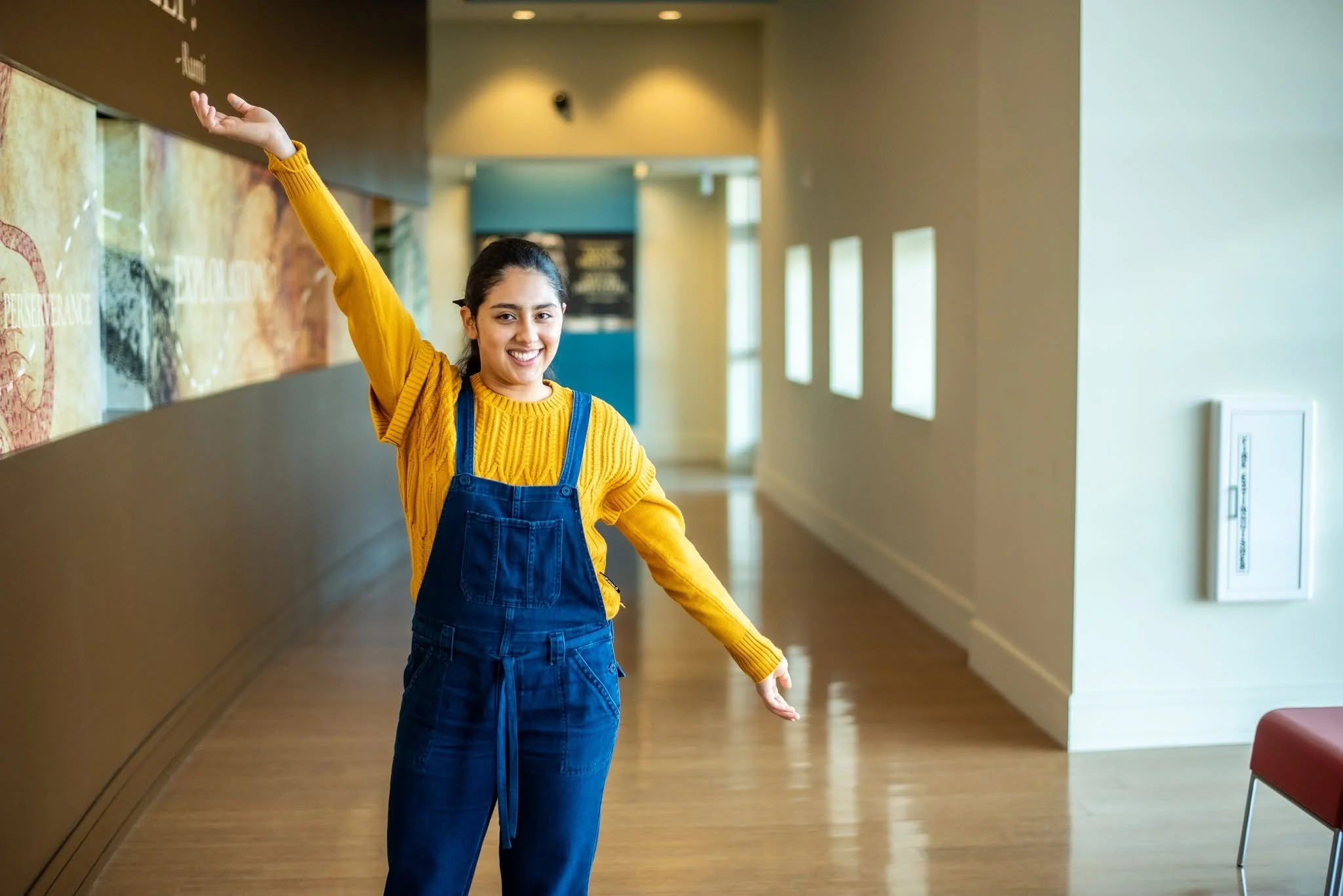 A young woman with dark hair tied back, smiling and waving her right hand, standing in a well-lit indoor space. She is wearing a yellow sweater and blue overalls. The background features a hallway with windows on the right and a wall with artistic displays on the left side.