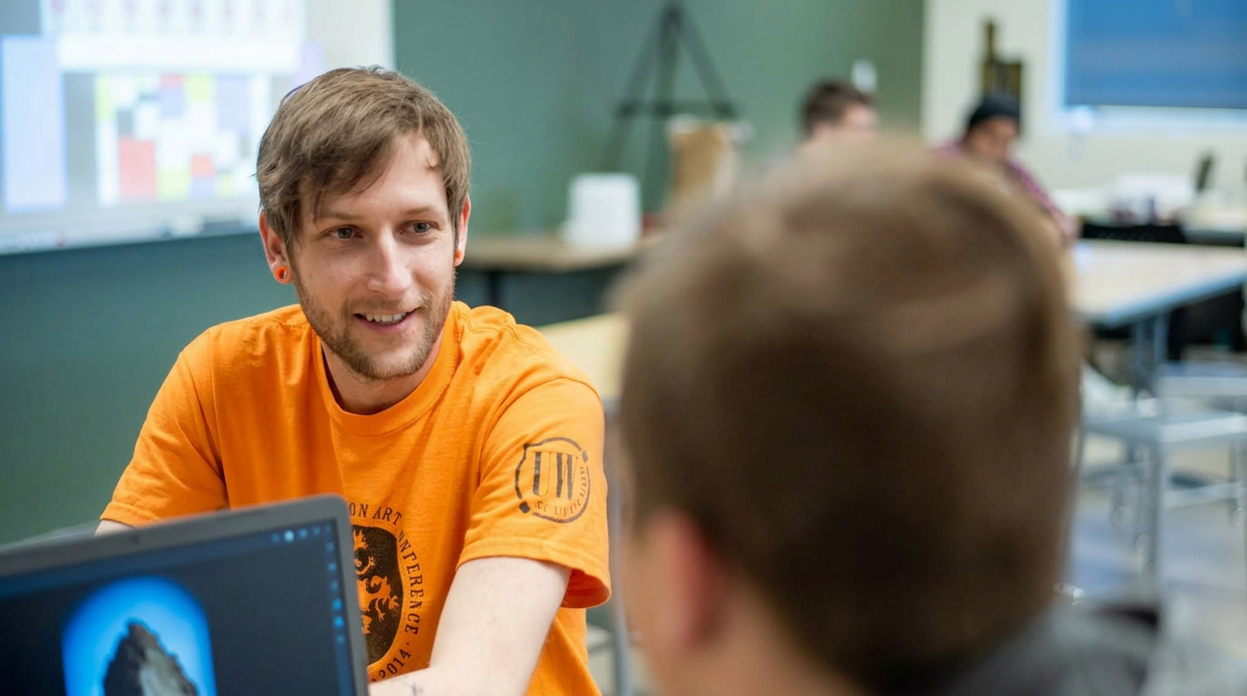 A person with short brown hair and a beard, wearing an orange T-shirt, is smiling and talking to someone with glasses in a classroom setting. Other students are visible in the background working at desks.