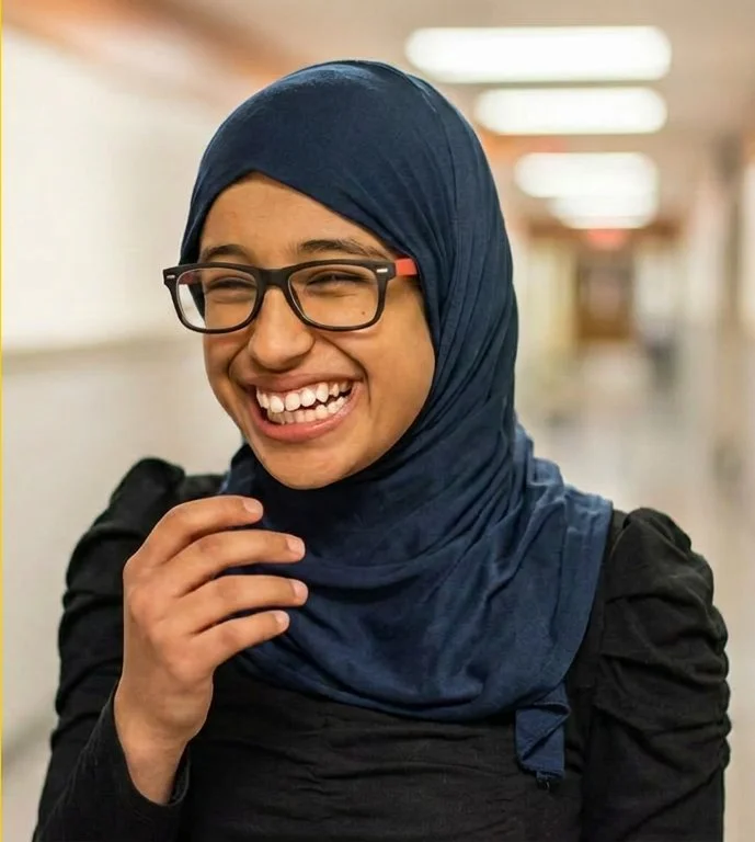 Young woman smiling, wearing glasses and a blue hijab, in an indoor setting.