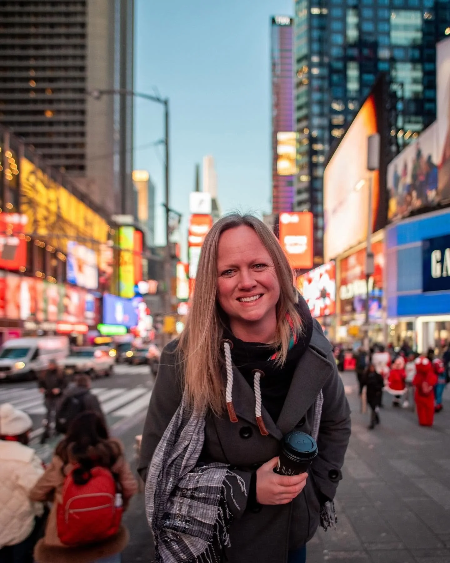 A woman smiling while holding a coffee cup in Times Square, New York City, during the evening with busy crowds and lit-up billboards in the background.