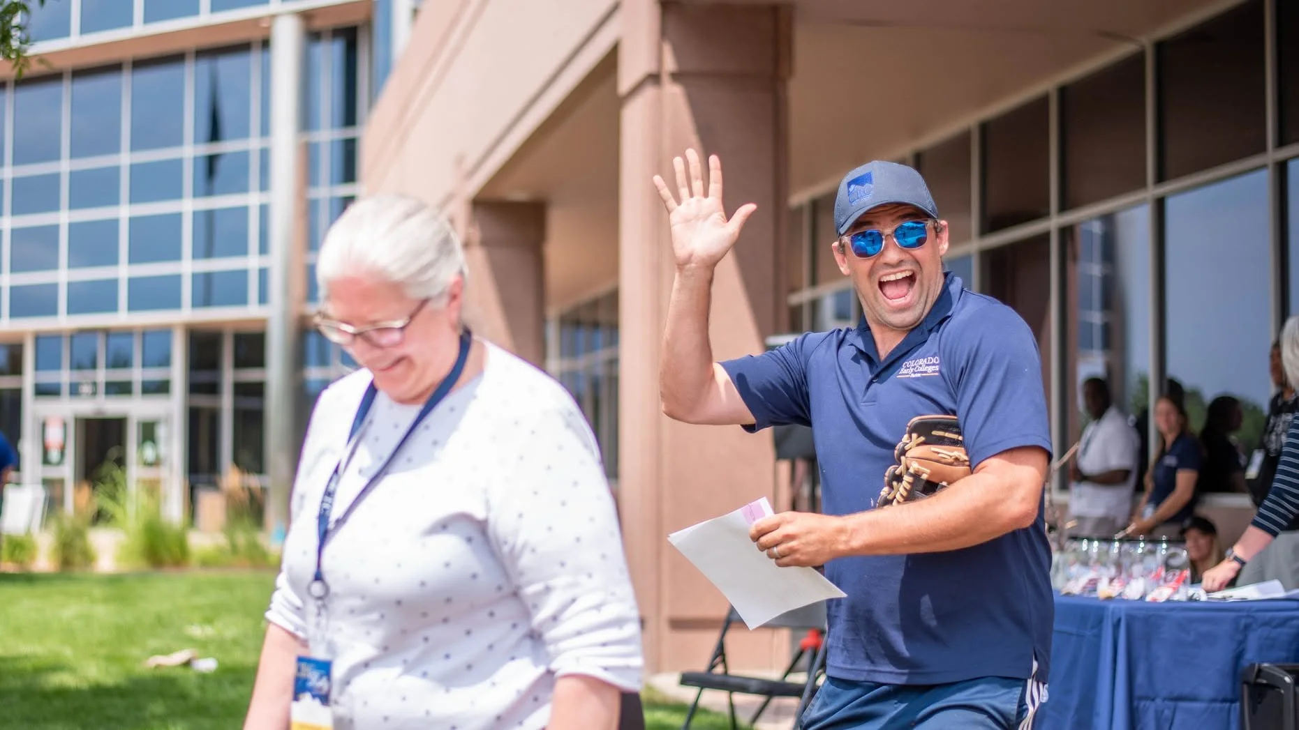 A cheerful man wearing sunglasses, a baseball cap, and a polo shirt waves and smiles outdoors at a community event, with a woman in front of him and other people and tables in the background.