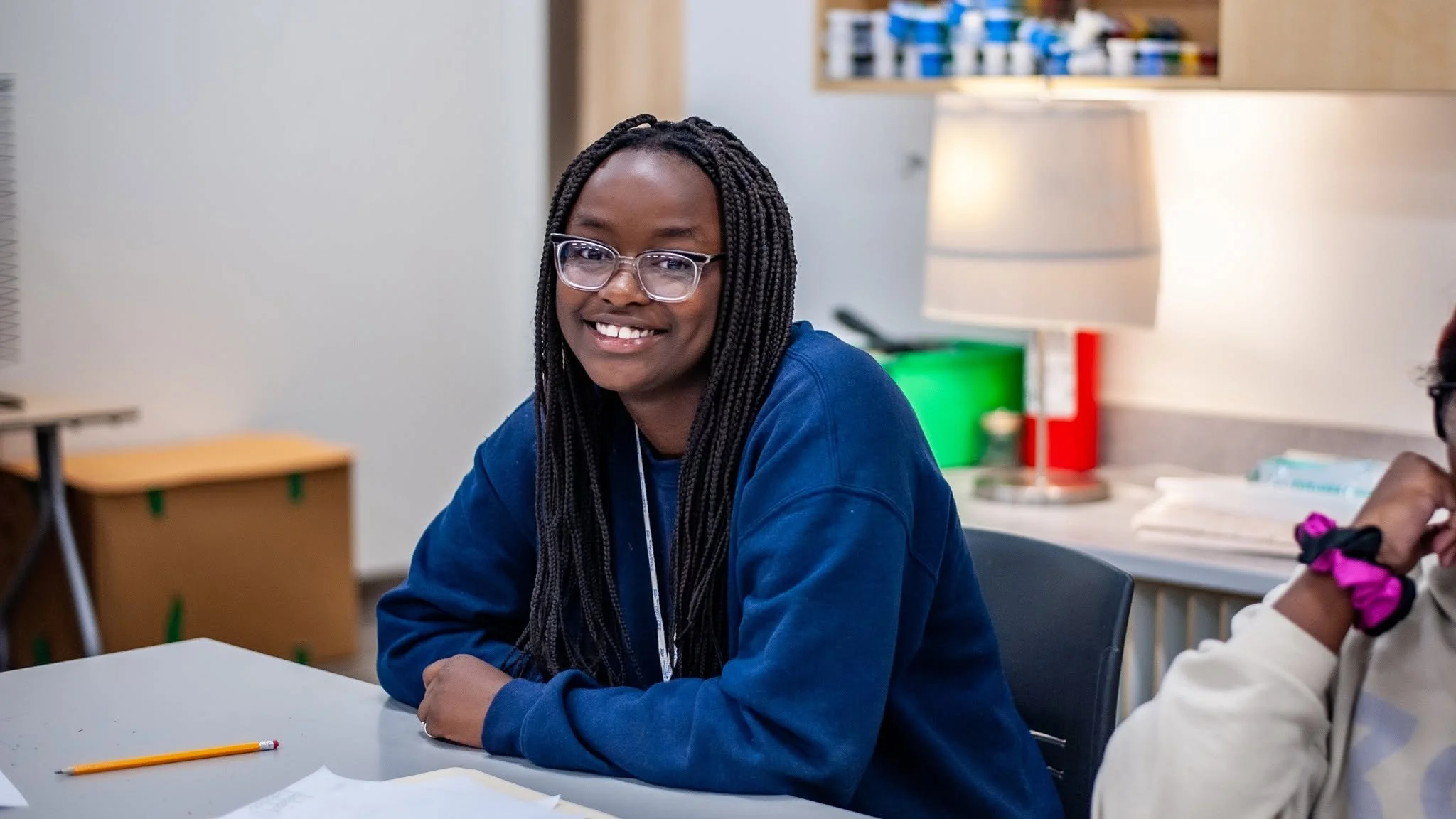 A young woman with long braided hair, wearing glasses and a blue hoodie, smiling while sitting at a table in a classroom or office setting with a desk, lamp, and shelves in the background.