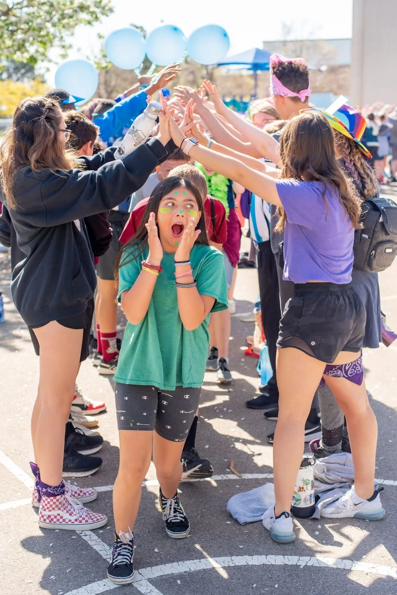 A group of children dressed in casual clothing and sneakers, participating in an outdoor event. They are standing in a circle, high-fiving each other, with some wearing hats and face paint. The setting appears to be a parking lot with balloons and decorations, and the children are smiling and happy.