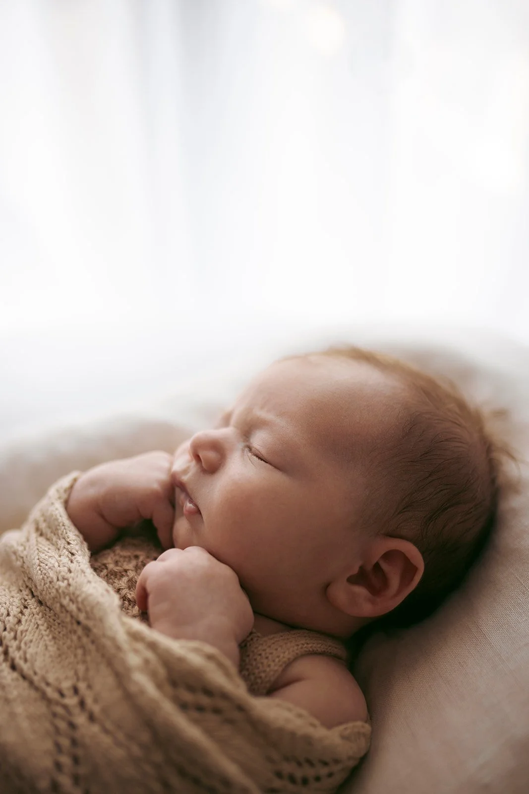 A sleeping newborn baby wrapped in a knitted blanket, lying on a soft surface, with closed eyes and relaxed facial expression.