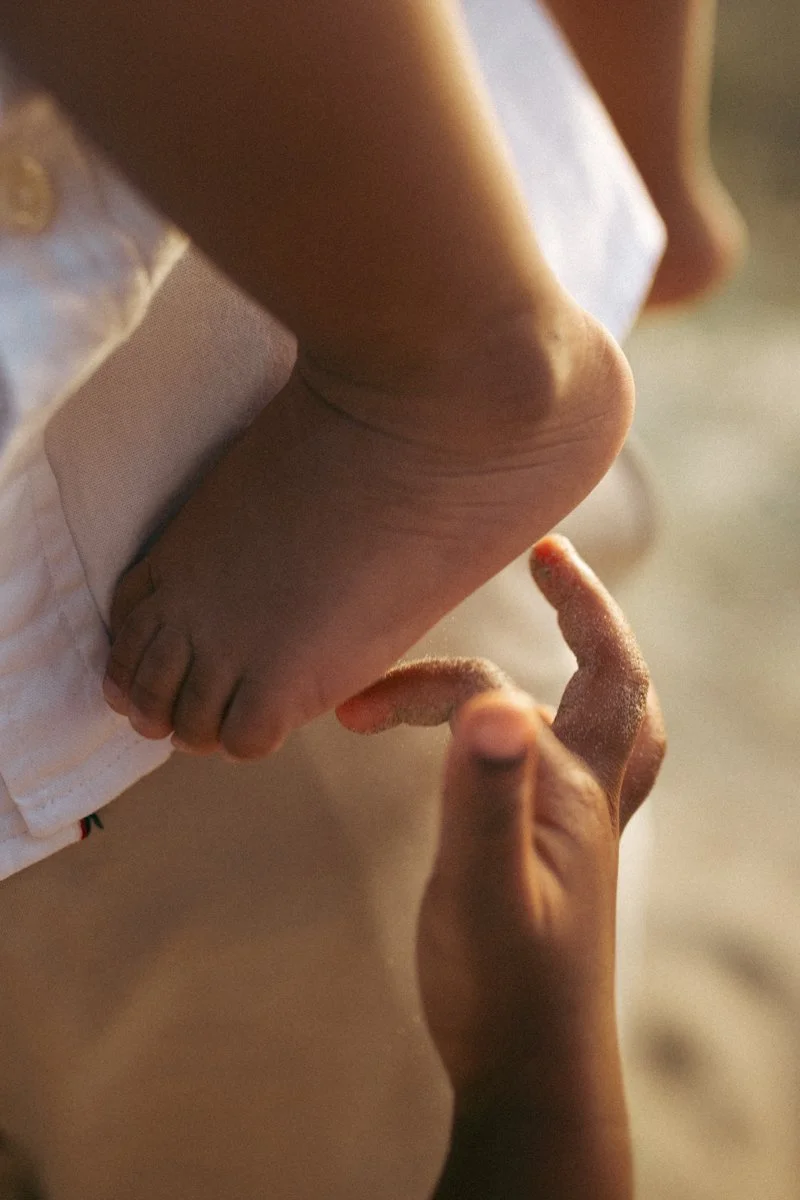 Close-up of a child's bare foot being touched by an adult's finger, with the child laying on a bed with white bedding.